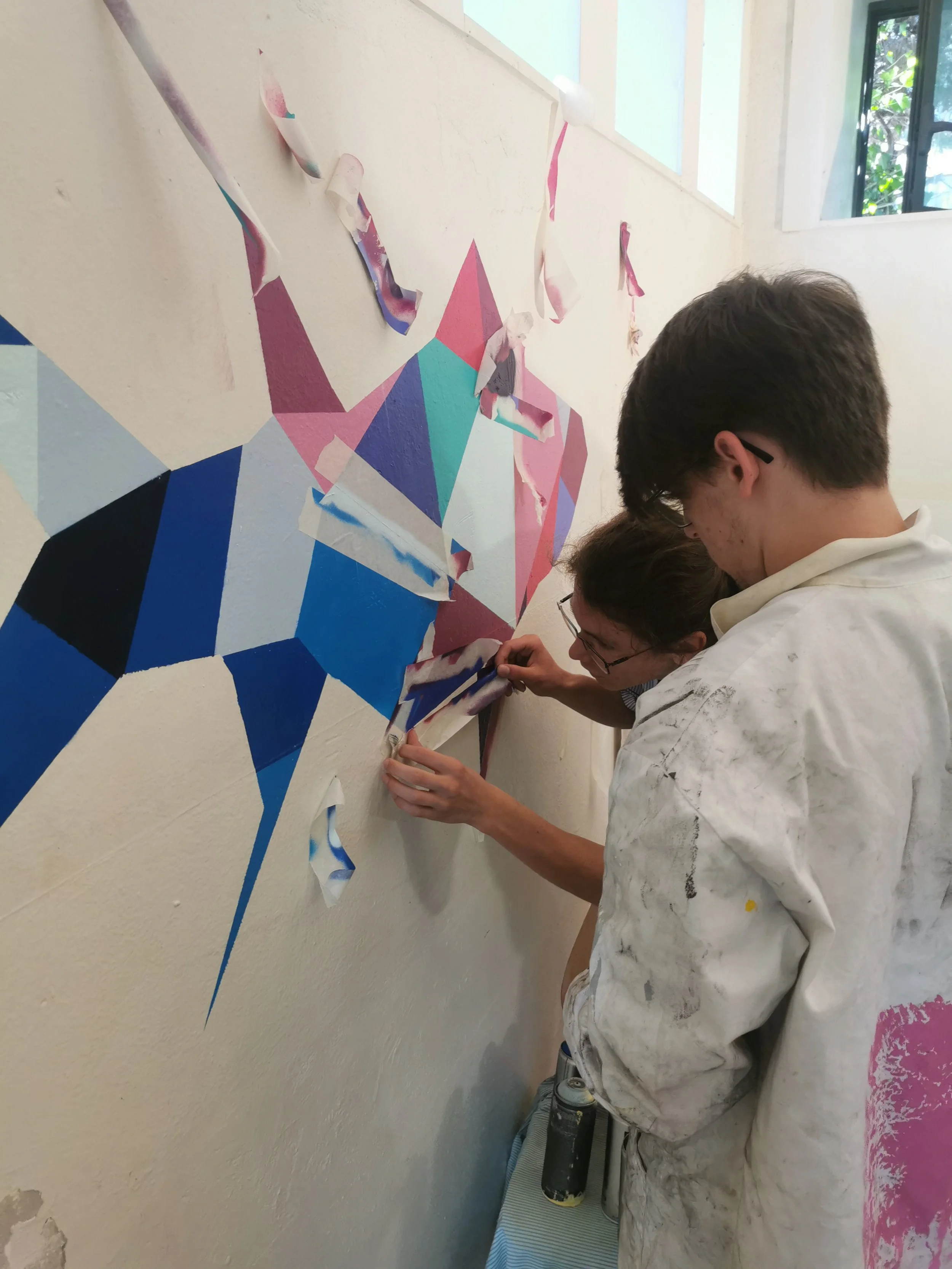 Deux jeunes hommes travaillent sur une fresque murale colorée dans une pièce lumineuse. Ils portent des vêtements de peinture tachés. EGGS EGGS58STUDIO