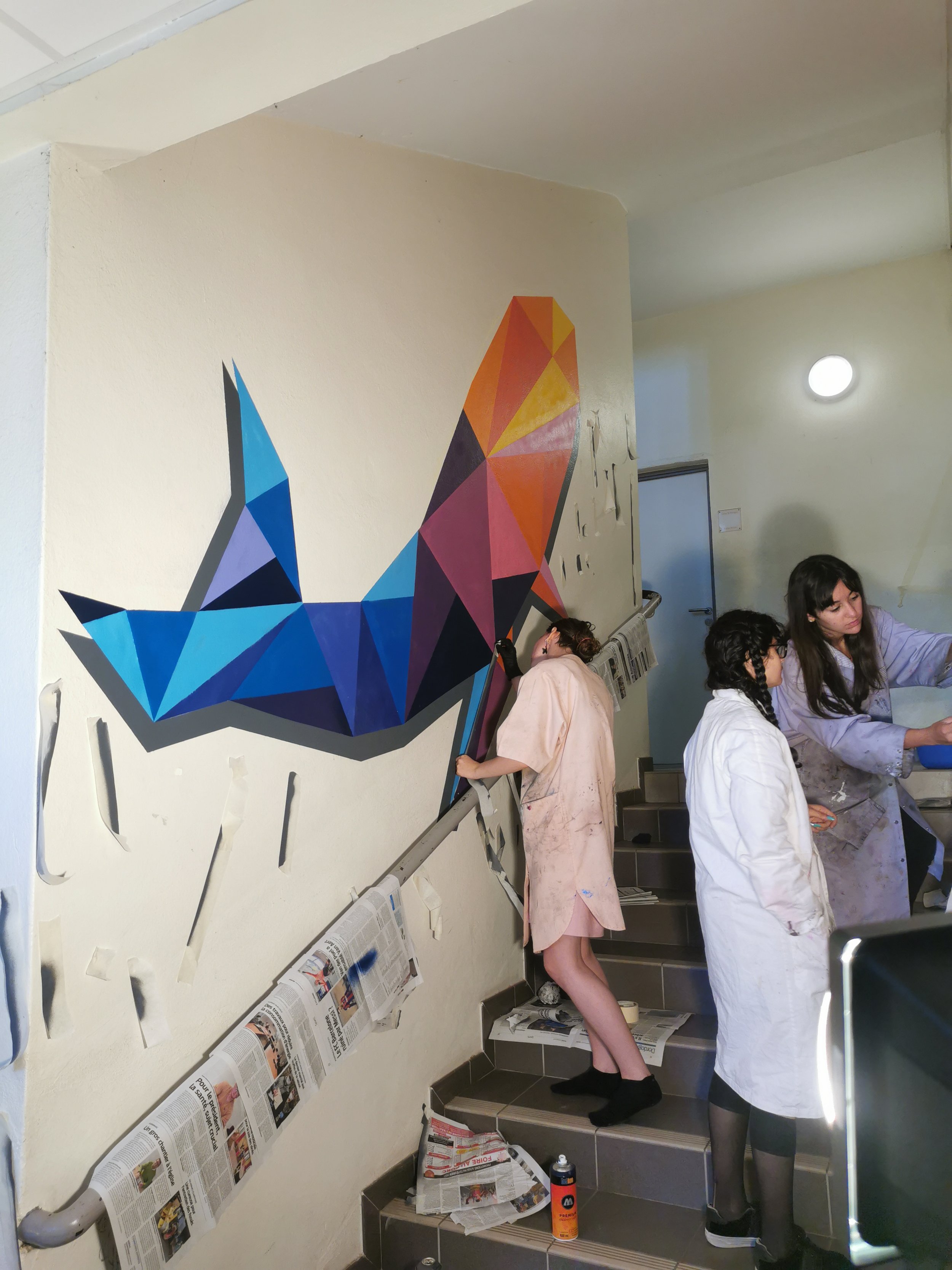 Groupe de personnes habillées en blanc et rose dans un espace intérieur, avec un mur décoré d'une peinture géométrique multicolore représentant un chien en forme polygonale. Certains regardent à proximité du mur, d'autres sont engagés dans une activi