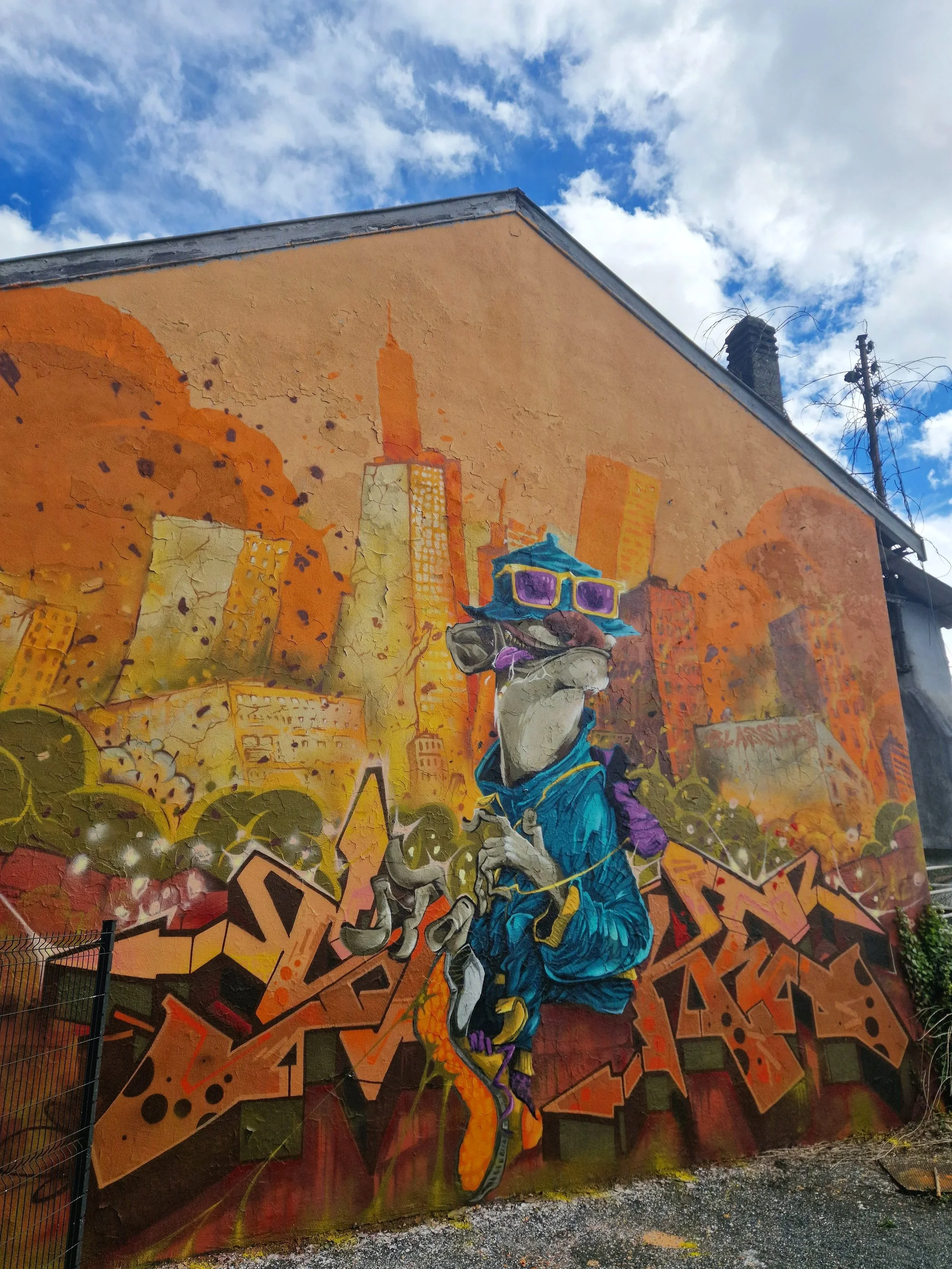 Une fresque murale colorée représentant un homme portant un casque et des lunettes de soleil, entouré de graffitis et de bâtiments en arrière-plan, sous un ciel bleu avec quelques nuages.