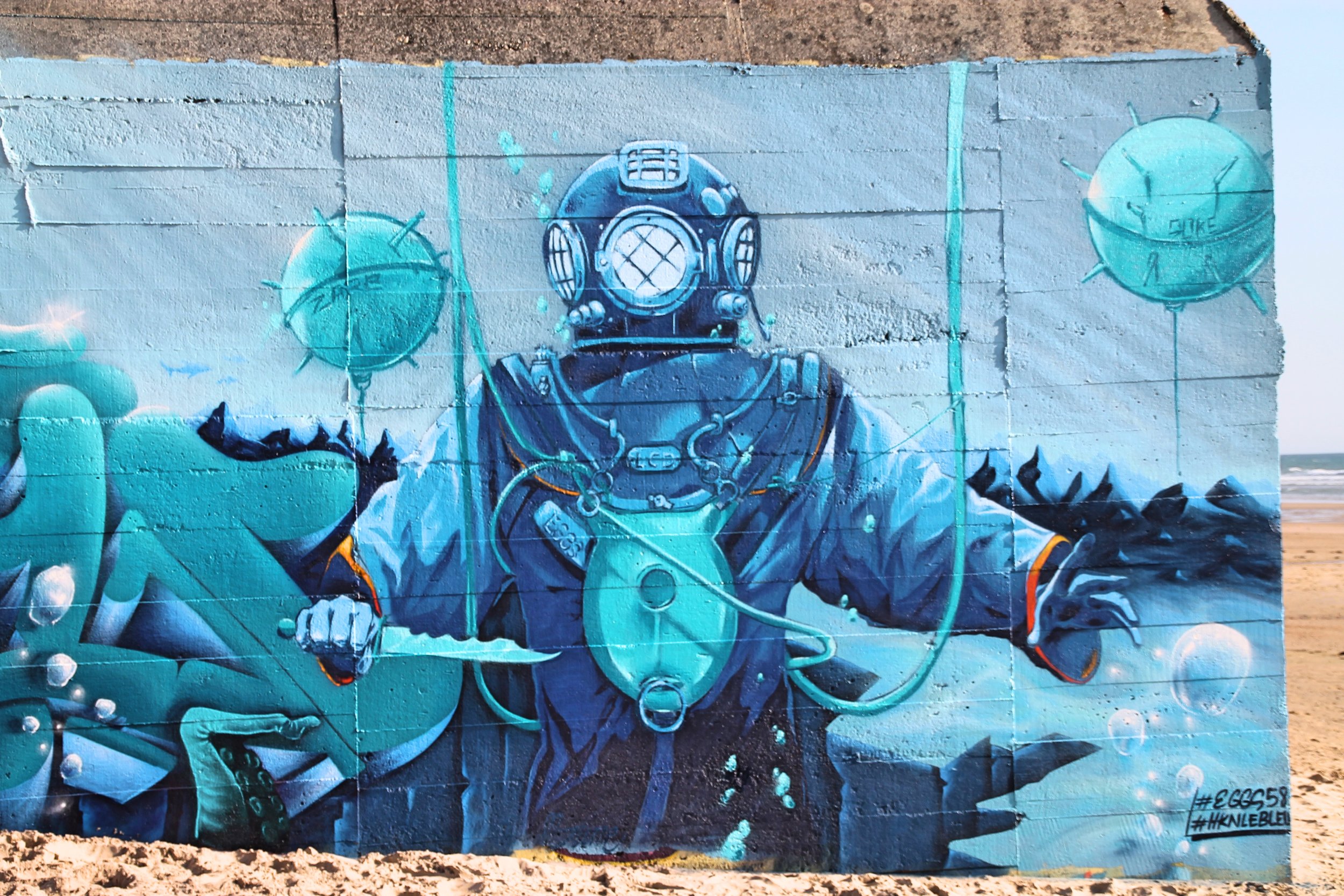 Une fresque murale représentant un plongeur en combinaison de plongée avec un masque à inhalation, entouré de bulles d'air et d'éléments marins, peinte sur un mur en dehors.