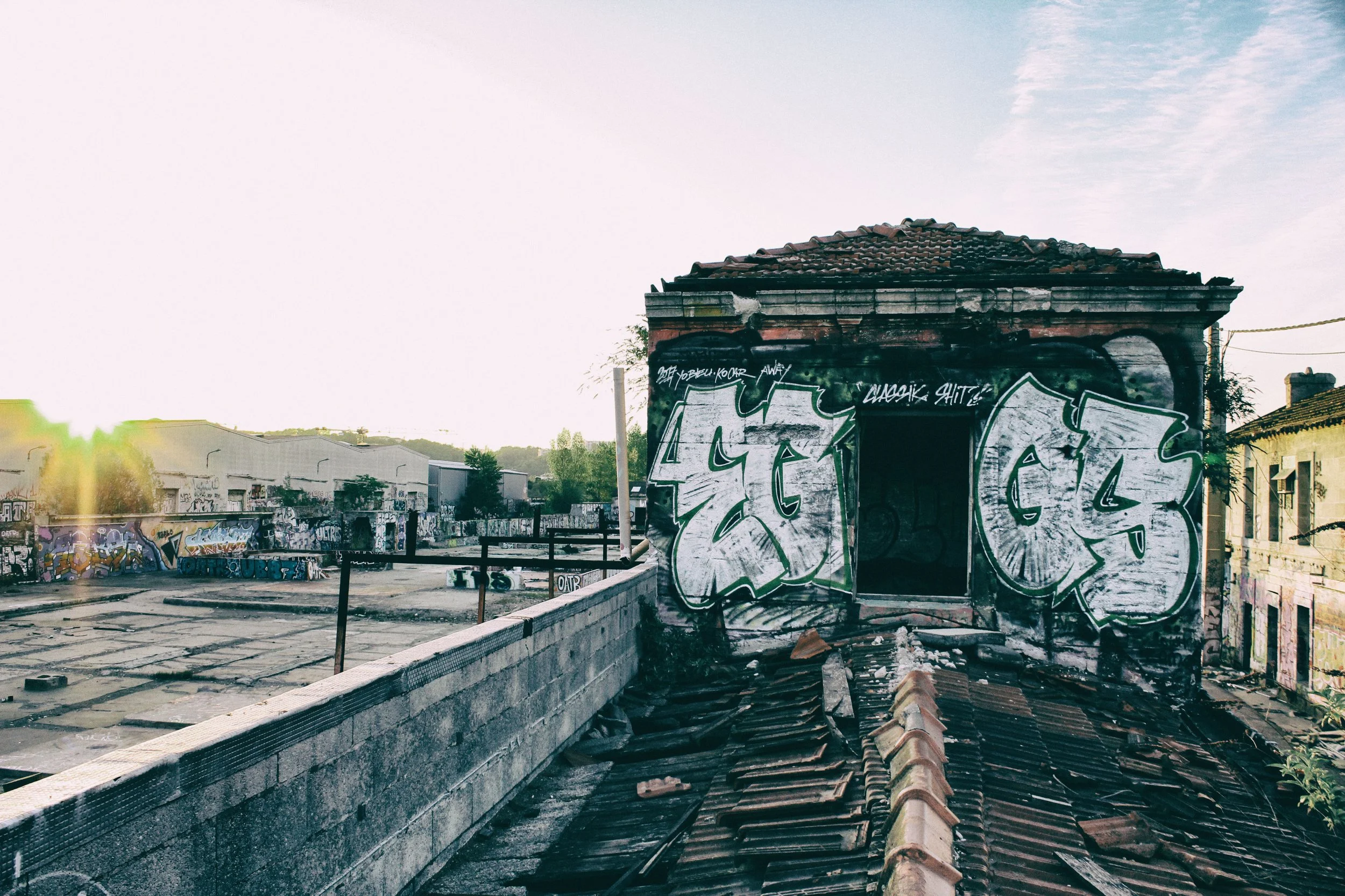 Un bâtiment abîmé avec des graffitis, un toit en tuiles cassé, dans un quartier en état de délabrement, avec une zone de construction ou de décharge à l'arrière-plan, sous un ciel en partie nuageux.