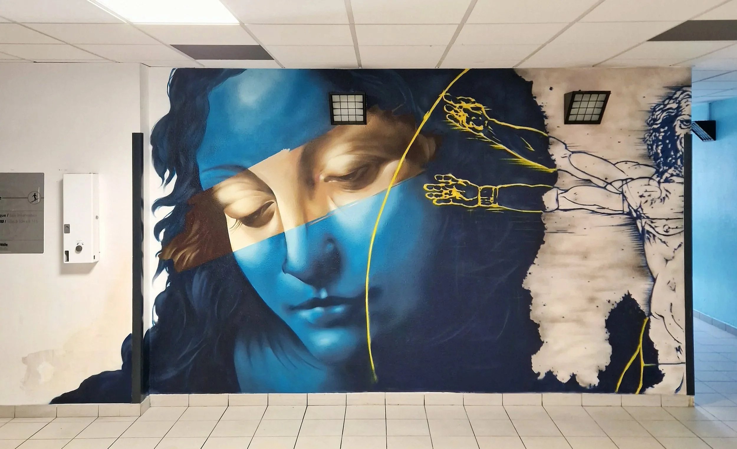 Une fresque murale représentant une face de femme, avec des éléments de peinture jaune et une pièce de puzzle en partie intégrée. La fresque est située dans un couloir avec un plafond en tuiles blanches et un sol en carreaux.