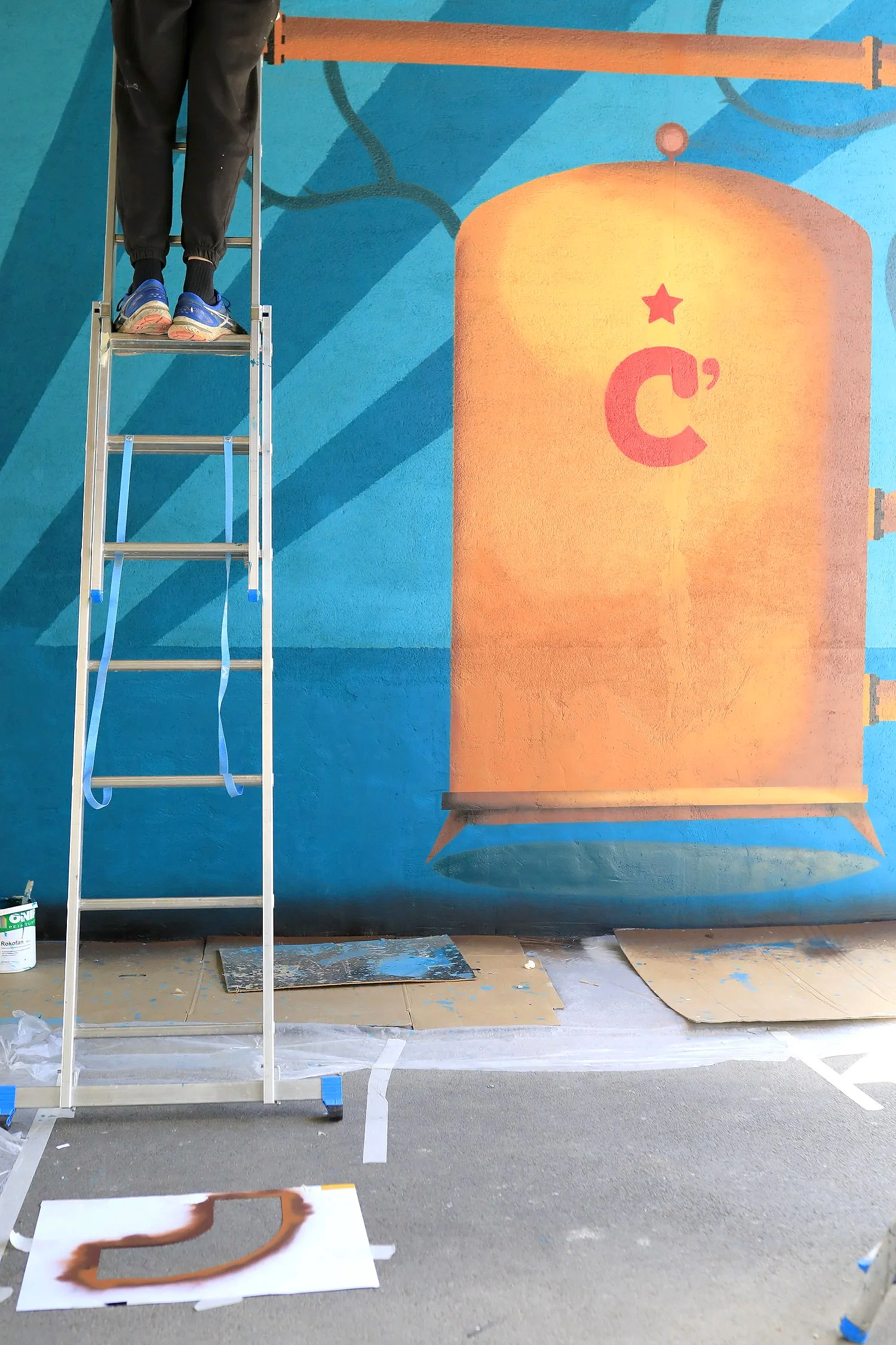 Un artiste peint un mur avec une fresque colorée, représentant une cuve a fermentation orange avec un logo en forme de C rouge (chavagn' brasseur) et une étoile rouge dessus, en utilisant une échelle et des outils de peinture.