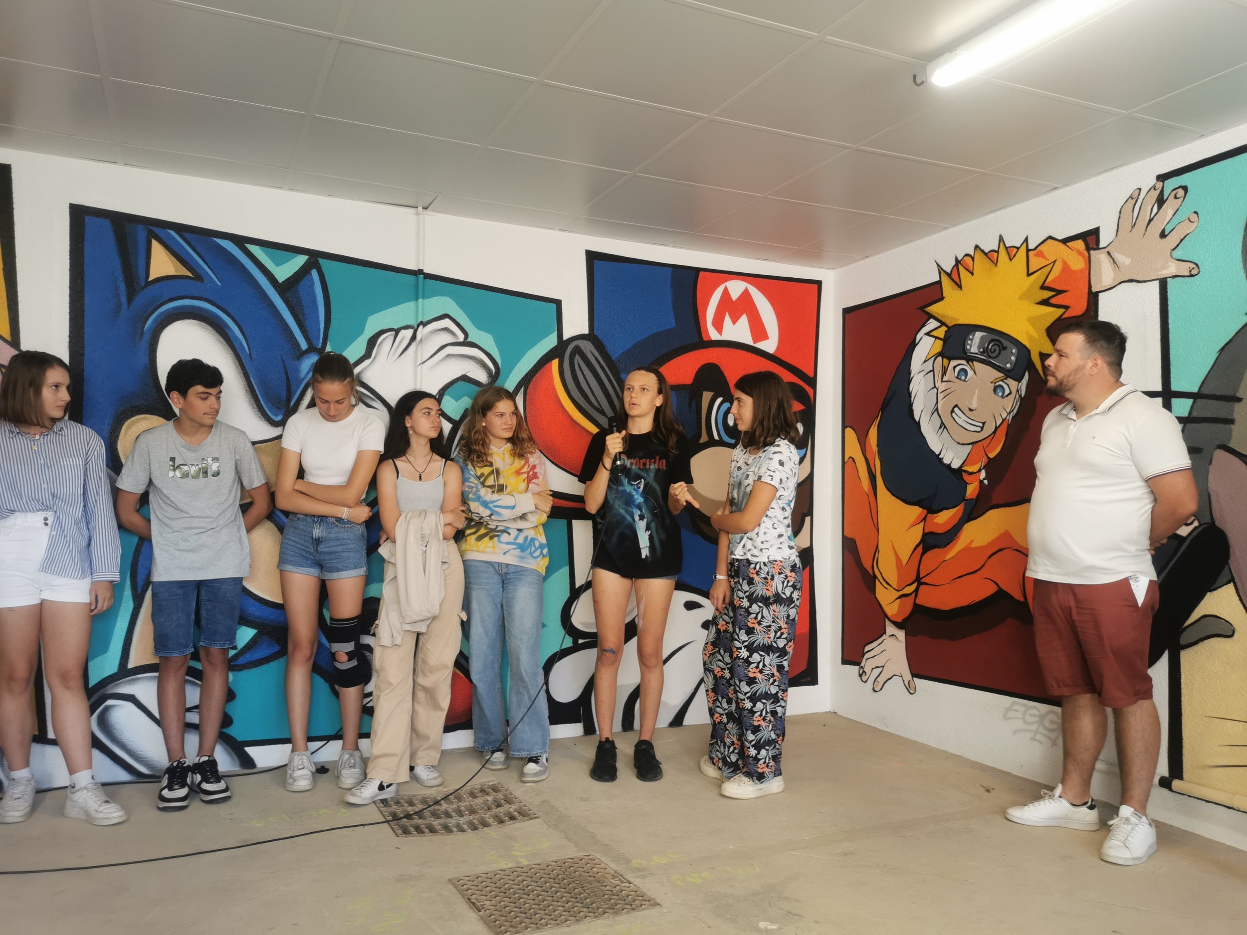 groupe de personne et un homme qui parlent dans une pièce avec des peintures à la sprays (street art) de dessins animés sur les murs.