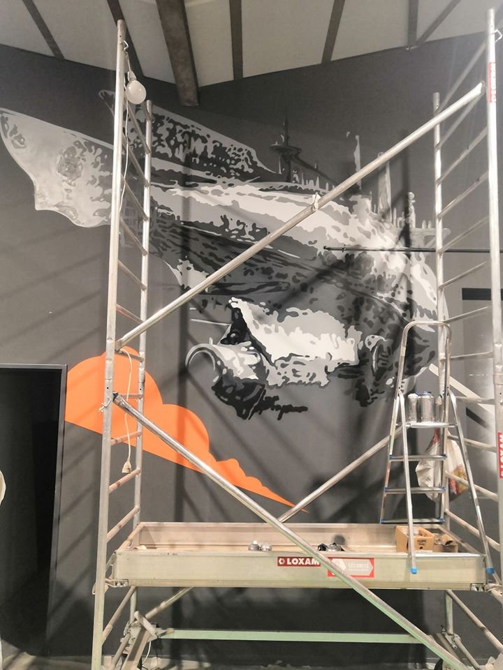 Mur en cours de peinture représentant un visage en noir et blanc avec une explosion orange au bas, échafaudage à côté.