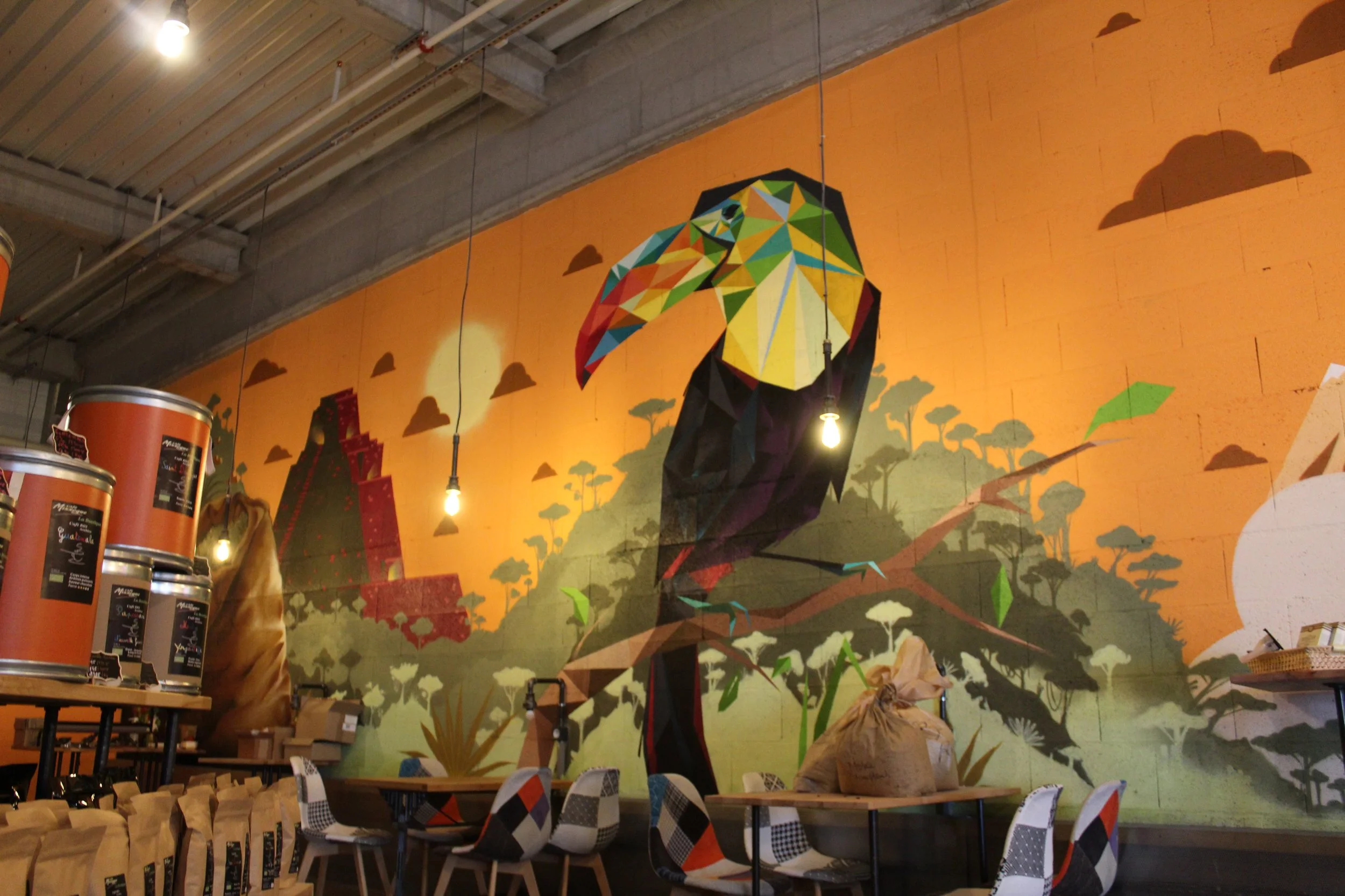 Mural coloré représentant un paon stylisé dans un intérieur de magasin ou café avec des tables et chaises en premier plan.