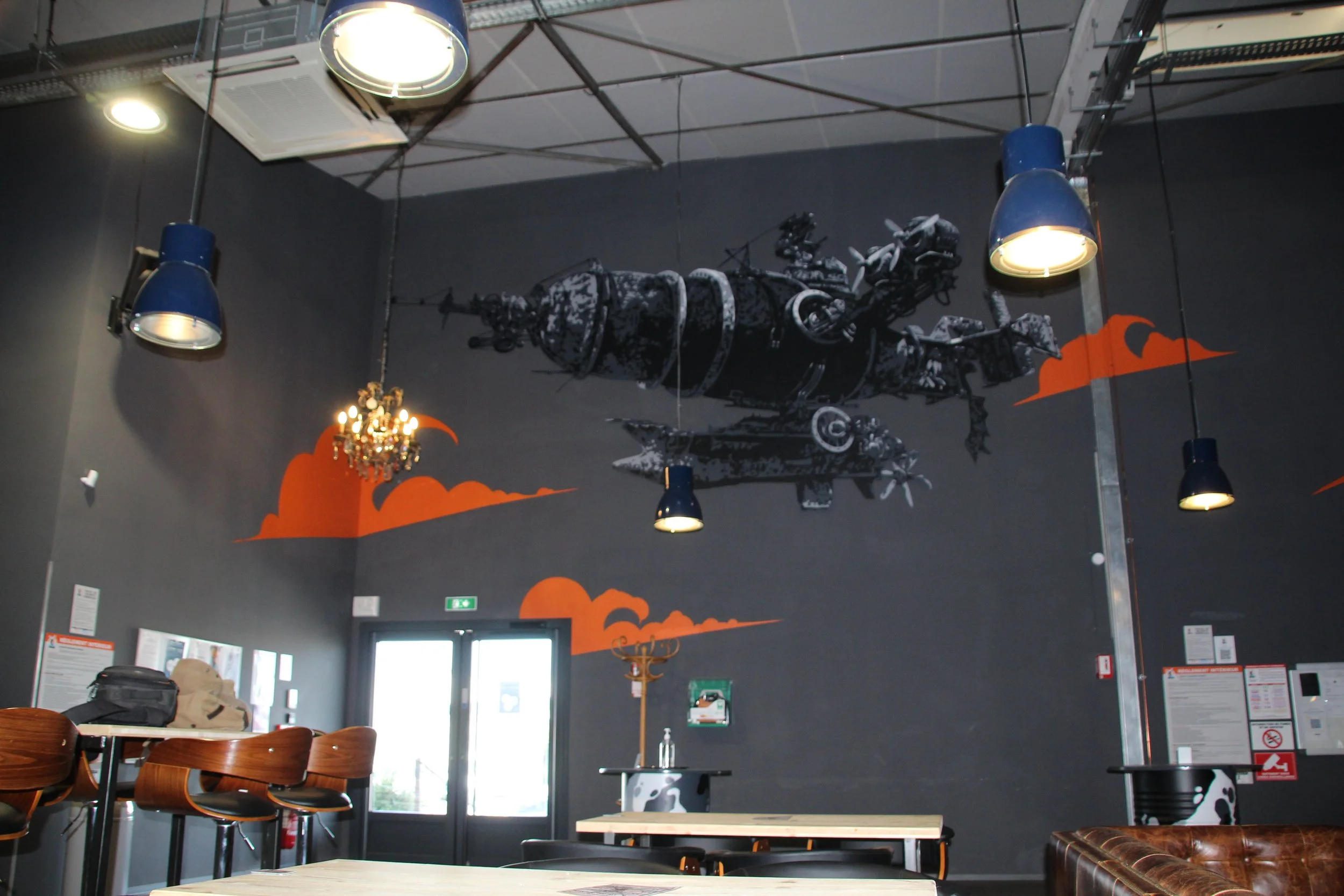 Intérieur d'un café ou d'un restaurant avec un décor industriel, une peinture d'une scène spatiale avec des drones ou des machines volantes, éclairage suspendu, tables et chaises en bois, une lampe à chandelier et un mur gris foncé avec des œuvres d'art simplifiées.