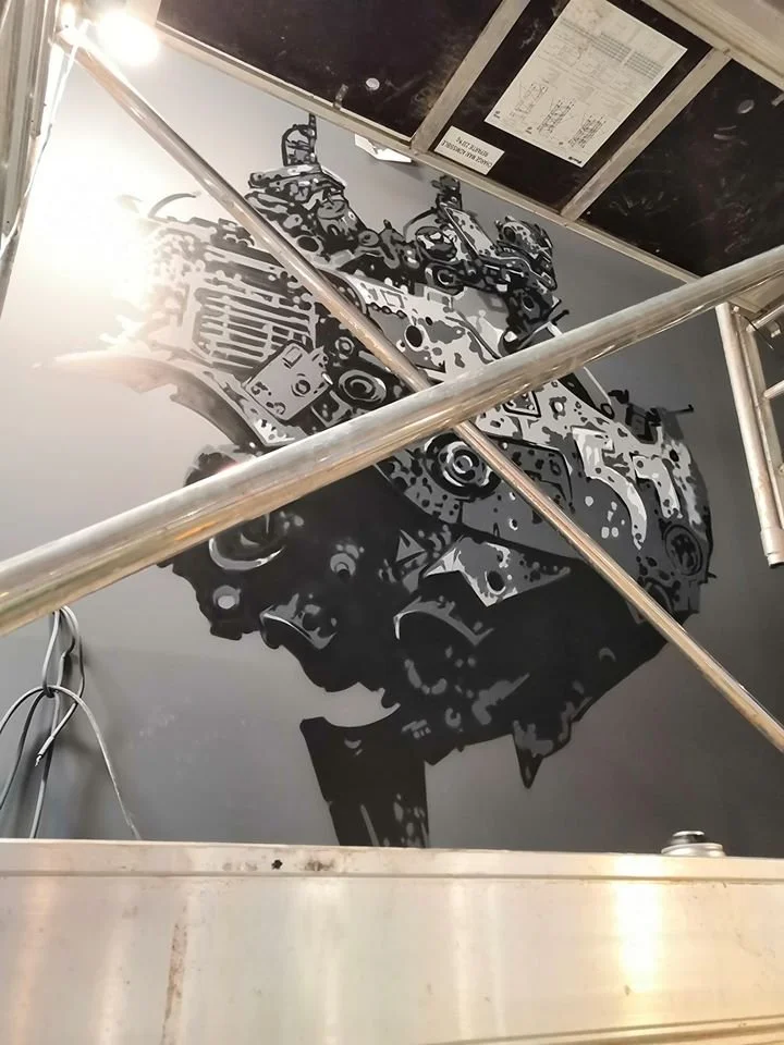 Une peinture murale en noir et blanc représentant un moteur d'automobile, vue du sol avec une échelle en aluminium dans un atelier ou un garage.