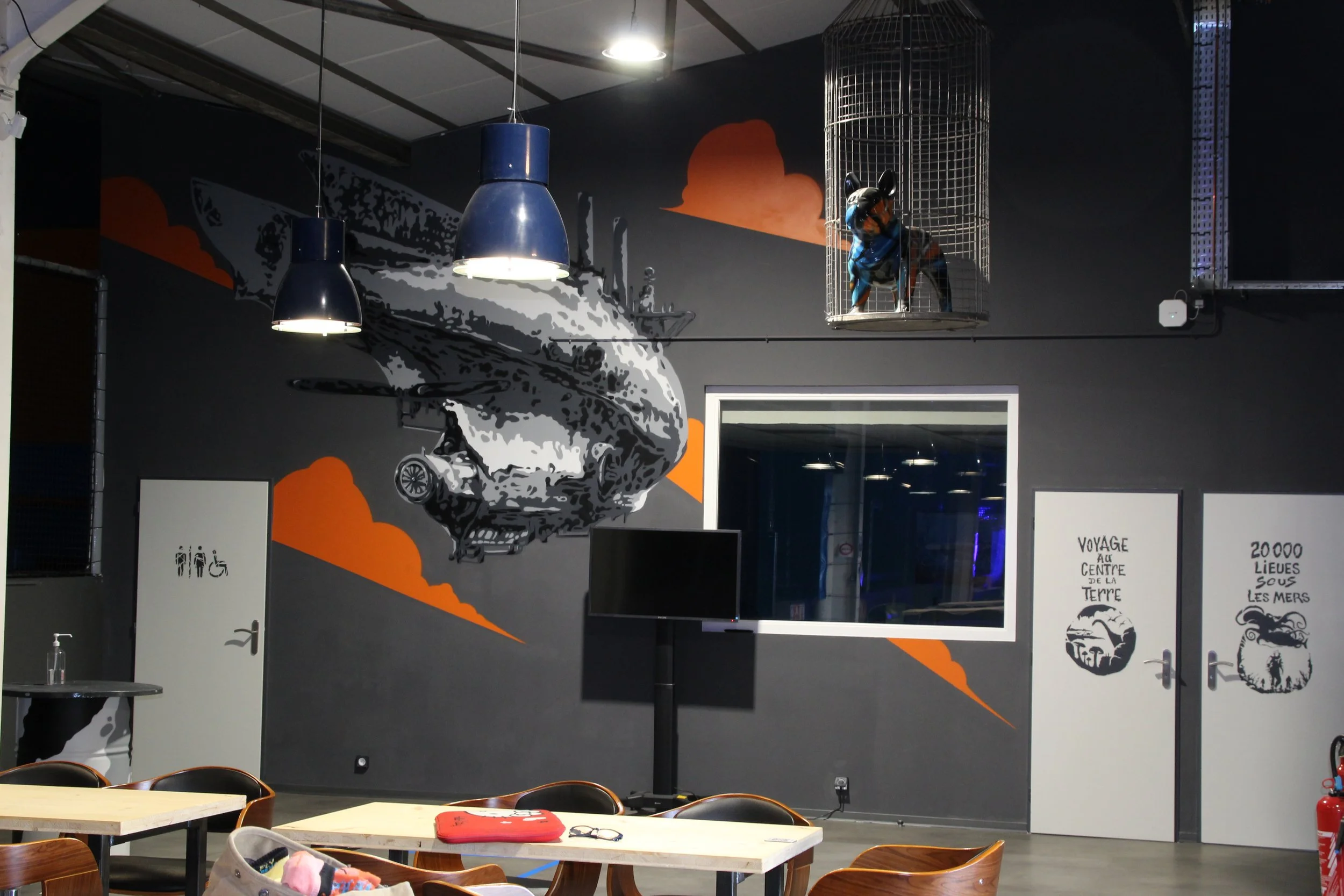 Une salle avec des tables, des chaises et un mur décoré d'une fresque représentant un avion de chasse avec des nuages orange et gris en arrière-plan. Une structure en cage avec un chien déguisé en pilote à l'intérieur est suspendue au plafond.