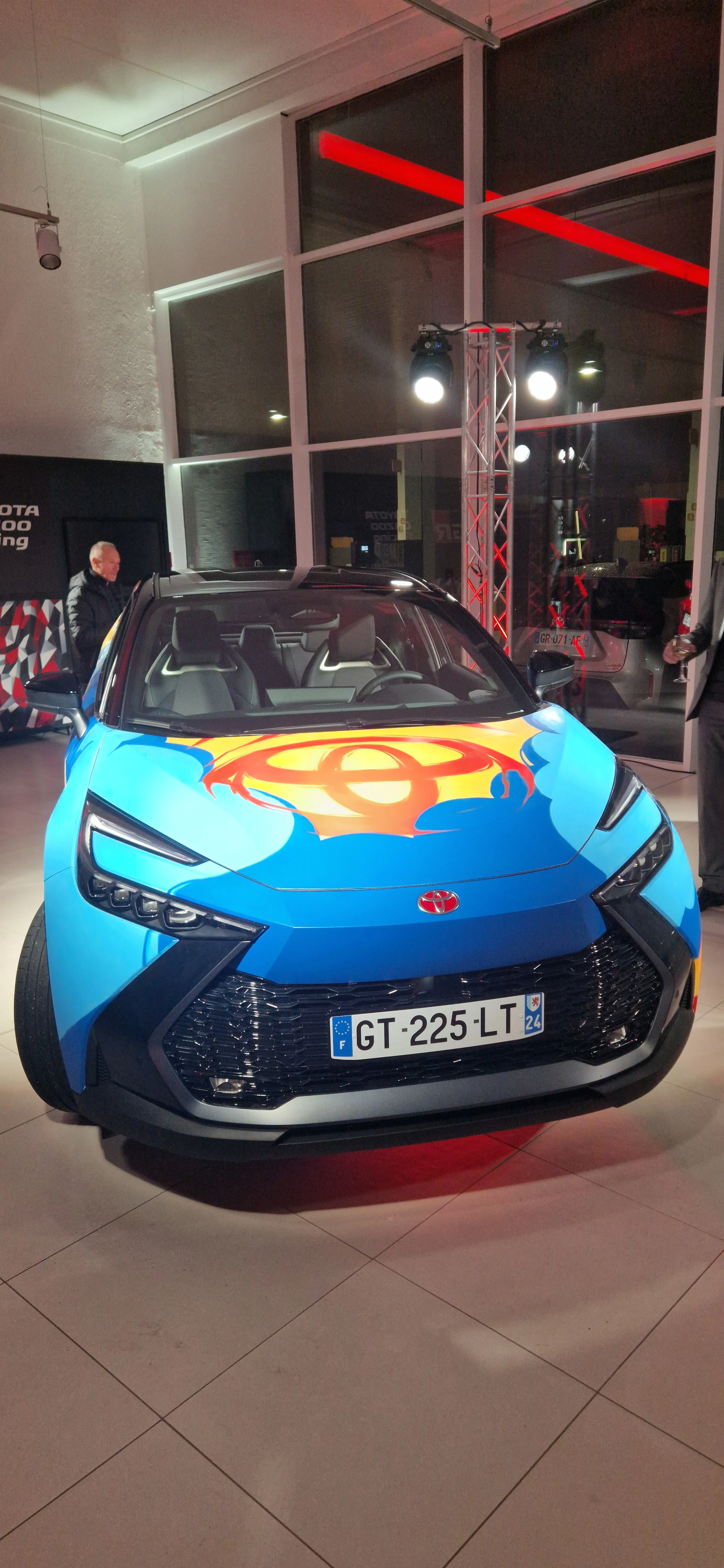 Une voiture électrique Toyota en couleur bleue avec un logo de Batman peint sur le capot, exposée à l'intérieur d'une salle avec des lumières d'éclairage intense et des vitres panoramiques.