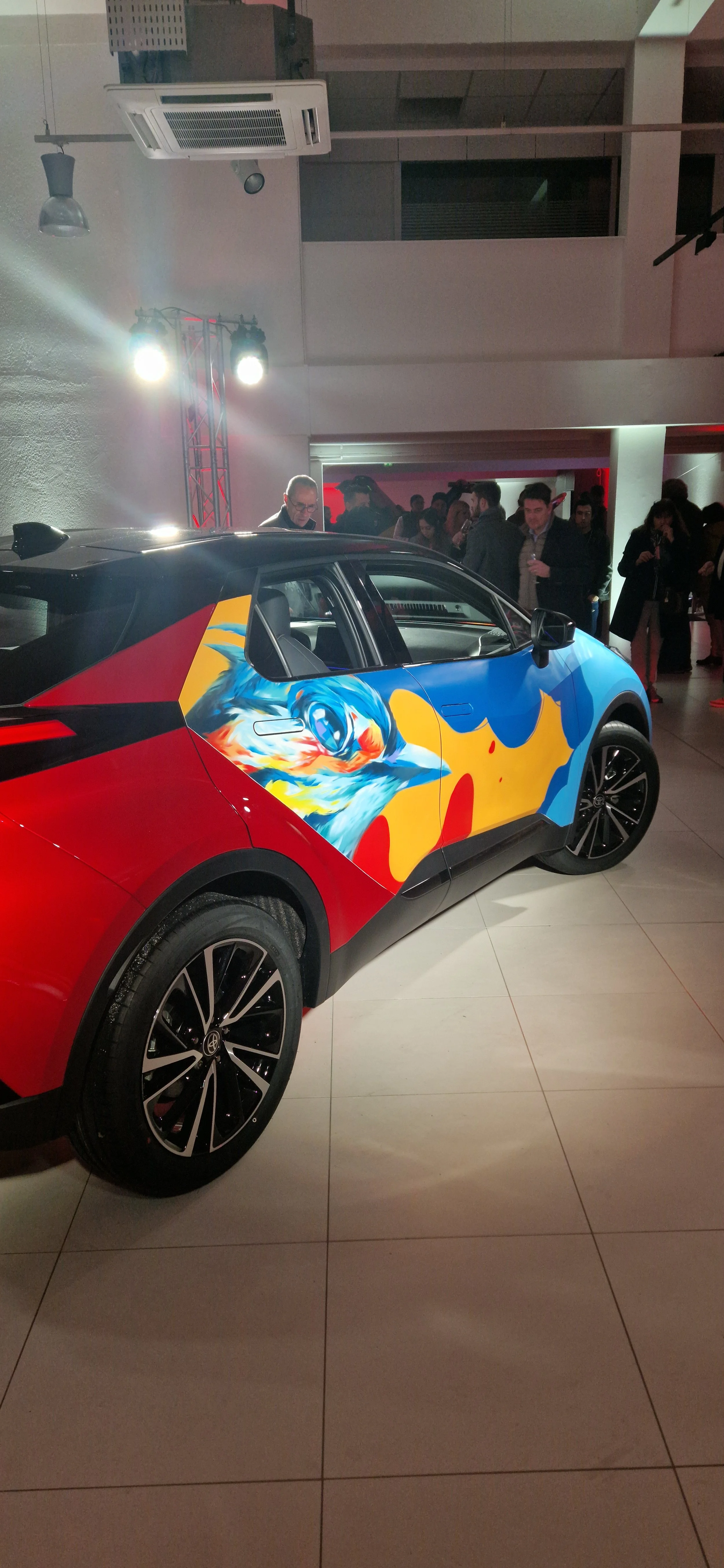 Voiture électrique de couleur avec un design coloré représentant un animal, probablement un chien, exposée lors d'un événement avec plusieurs personnes autour.