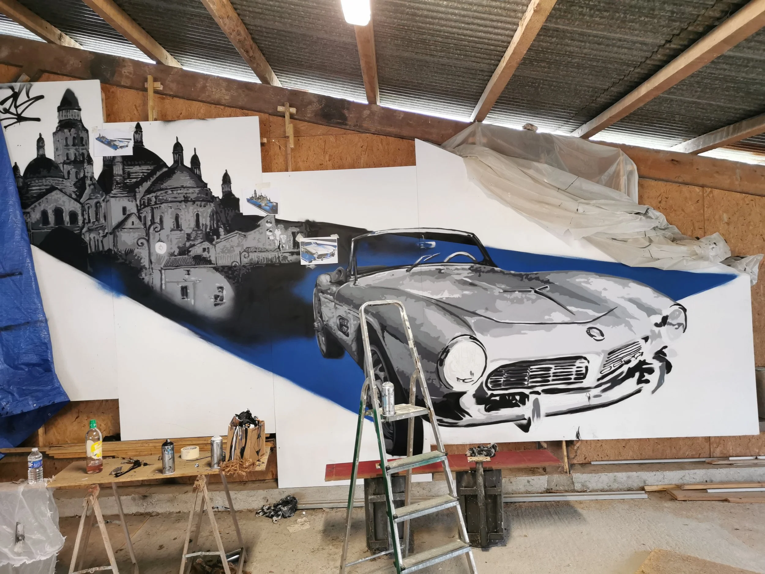Une grande peinture représentant une voiture de sport devant une vue d'une ville historique avec un bâtiment religieux. La scène est en noir et blanc avec des touches de bleu. La peinture est en cours de réalisation dans un atelier avec des outils et des bouteilles d'eau sur un tapis de sol, et un escabeau.