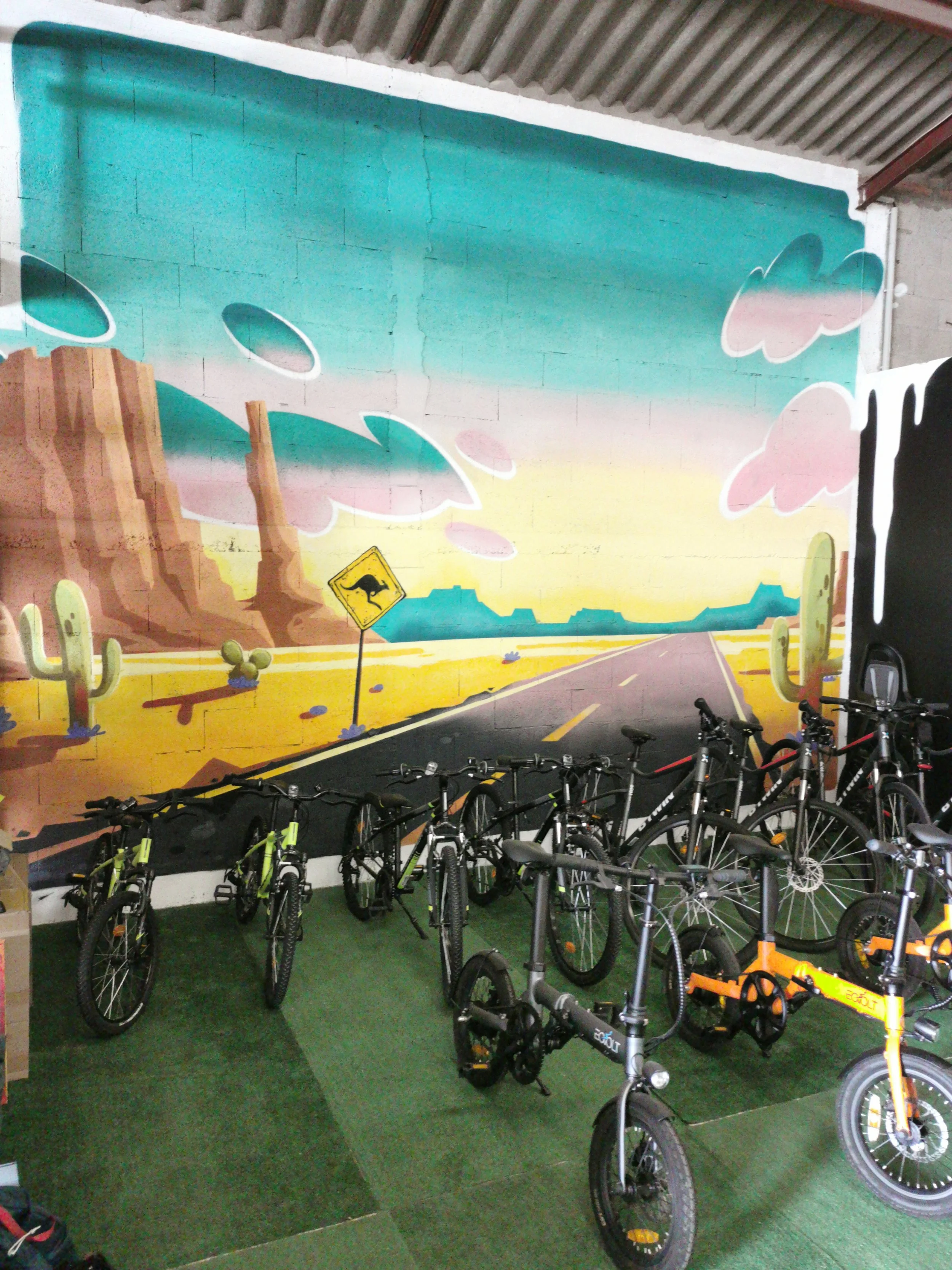 Vélos électriques rangés dans un espace couvert avec un mural coloré représentant un paysage désertique avec cactus, rochers et nuages roses, et un panneau de signalisation de kangourou.