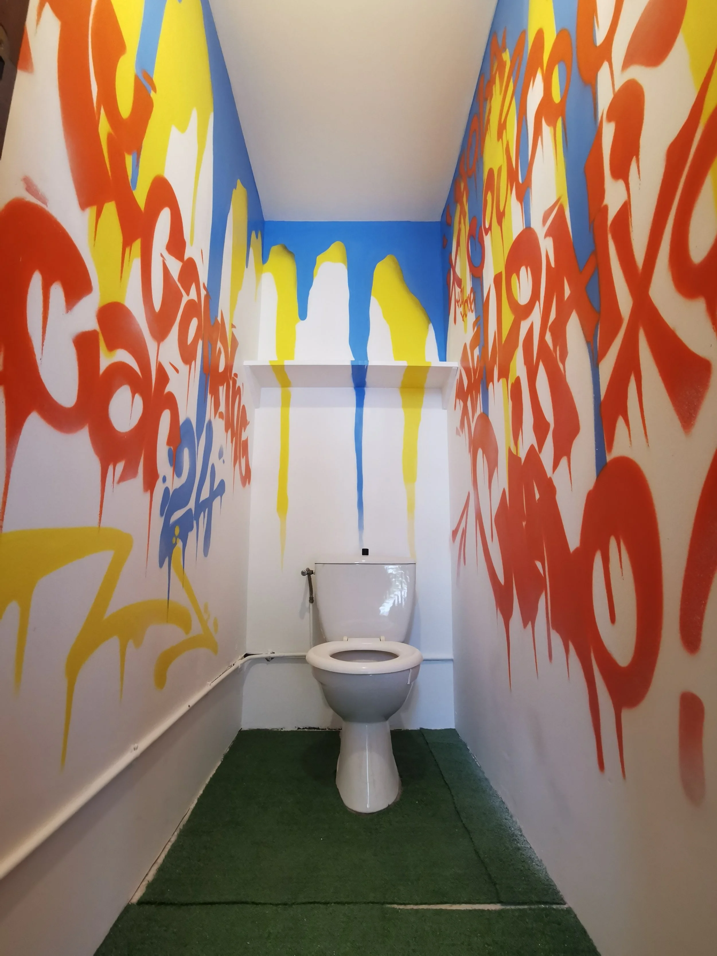 Toilettes dans une petite pièce avec des murs peints de graffiti coloré rouge, jaune, et bleu. Le sol est recouvert de moquette verte.