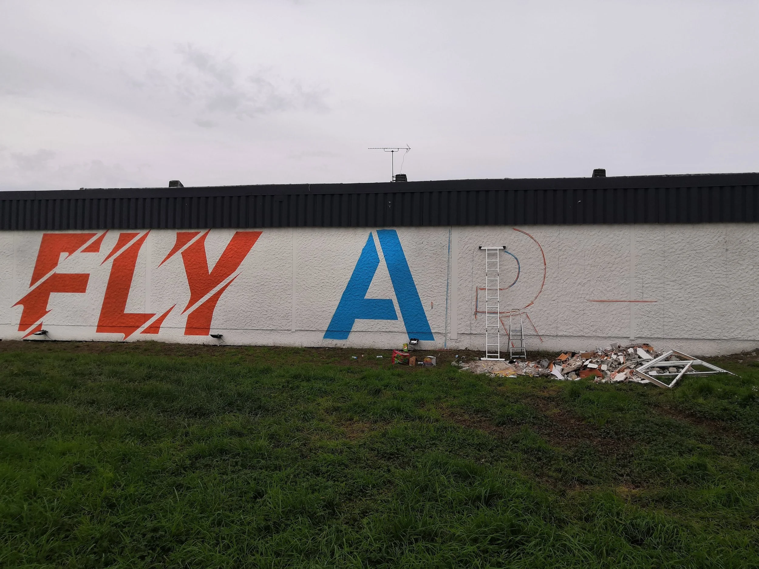 Une façade de magasin en cours de peinture avec des lettres en cours d'installation, un escabeau, des débris au sol, et un ciel nuageux au-dessus.