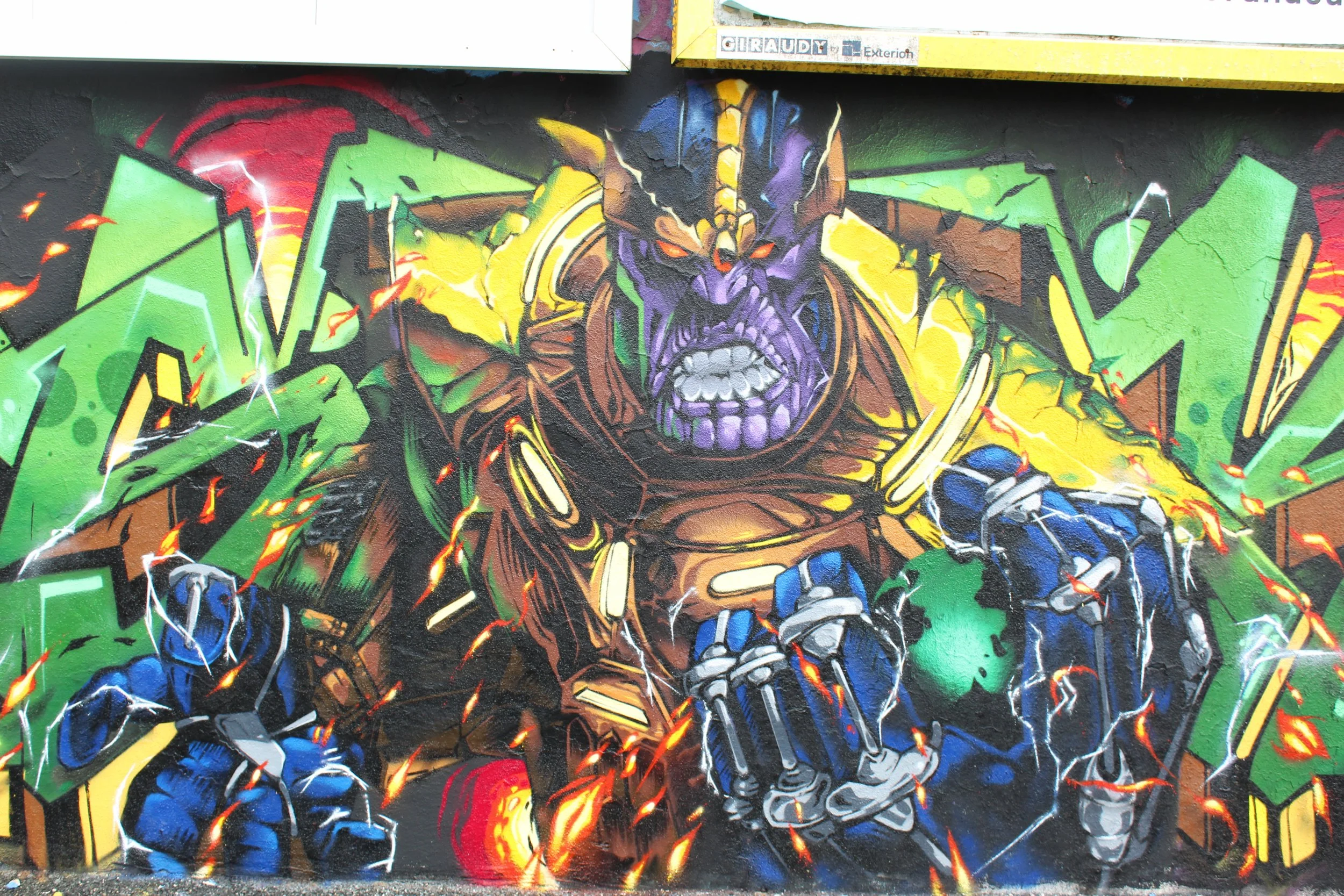 Graffiti de un personnage de comic avec un casque et un visage menaçant, coloré en violet, entouré de motifs verts, jaunes et bleus, avec des éclats de lumière et des fureurs de feu en arrière-plan.