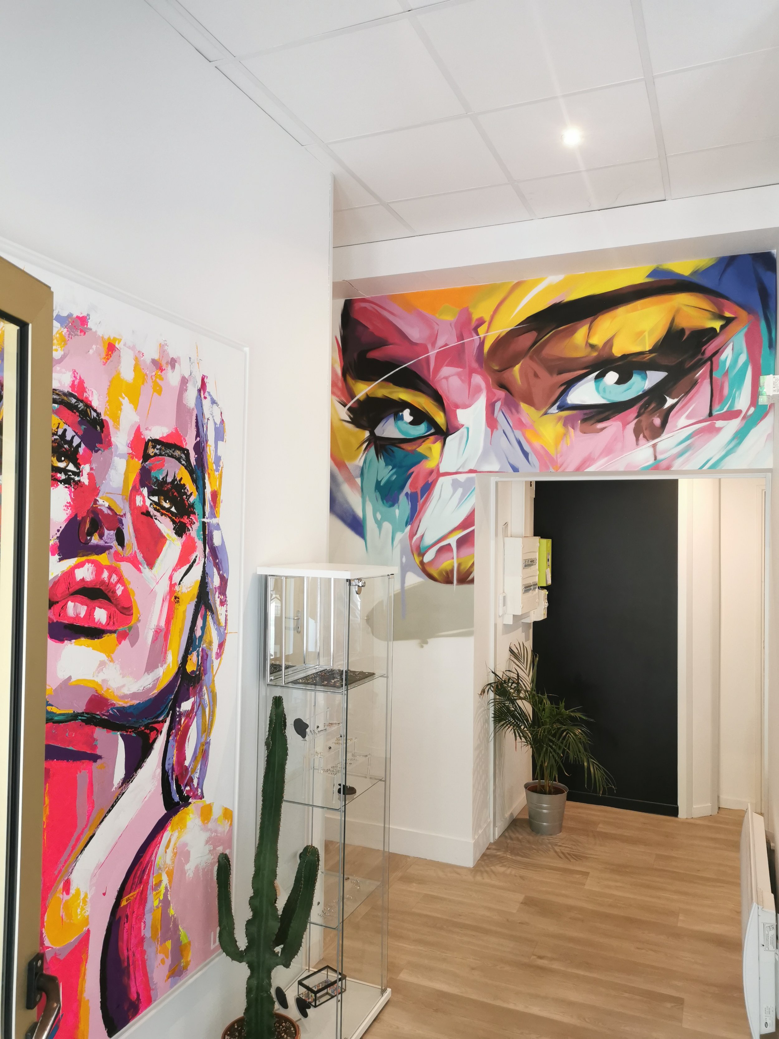 Une galerie d'art avec des peintures colorées, notamment un portrait de femme et un gros plan d'une autre. Il y a aussi une plante verte dans un pot et une étagère en verre avec des objets décoratifs.