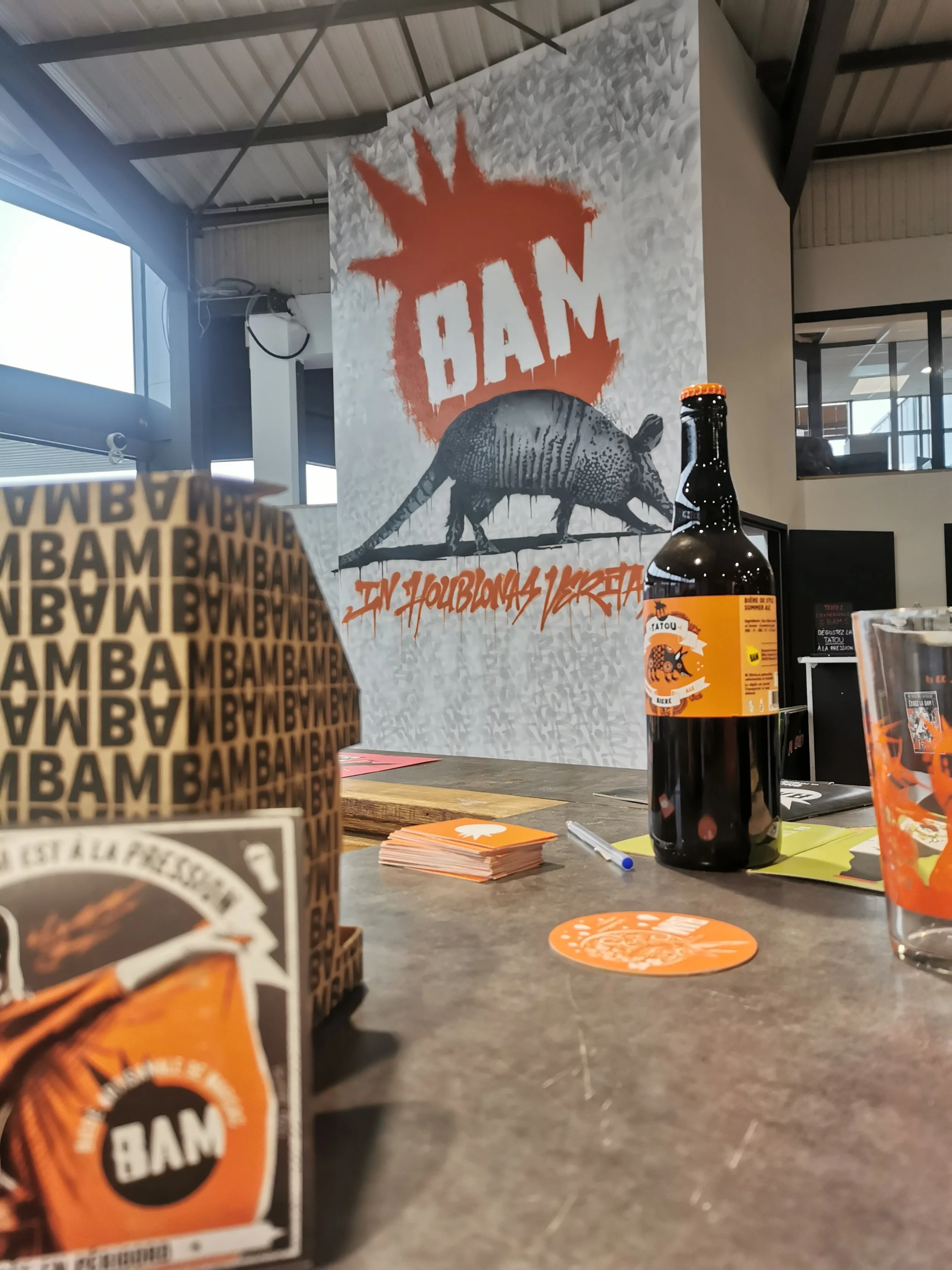 Une table avec divers objets promotionnels, une bouteille de bière, des flyers, au fond un affiche portant le nom 'BAM' avec une image d'un rat et le slogan 'En houblons Viretta'.