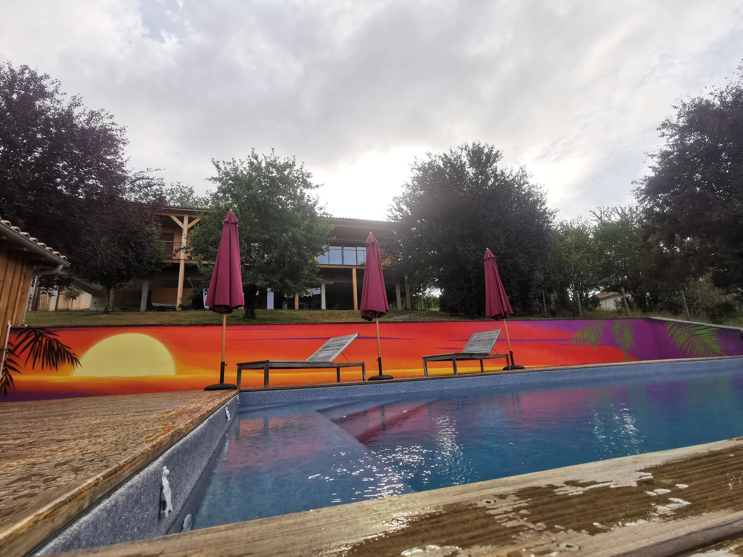 Piscine avec des parasols roses et une fresque colorée au fond, entourée d'arbres et d'une maison en bois. Ciel nuageux.