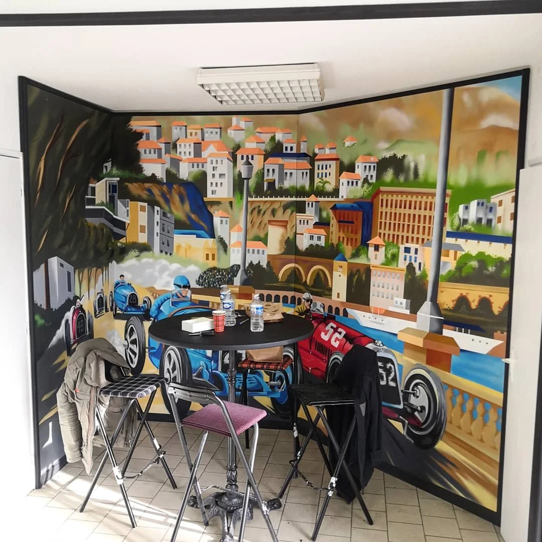 Un mur peint représentant une scène de course automobile avec des voitures de course colorées, un paysage urbain avec des immeubles et des ponts, et une cascade, dans une pièce avec une table ronde et des chaises.