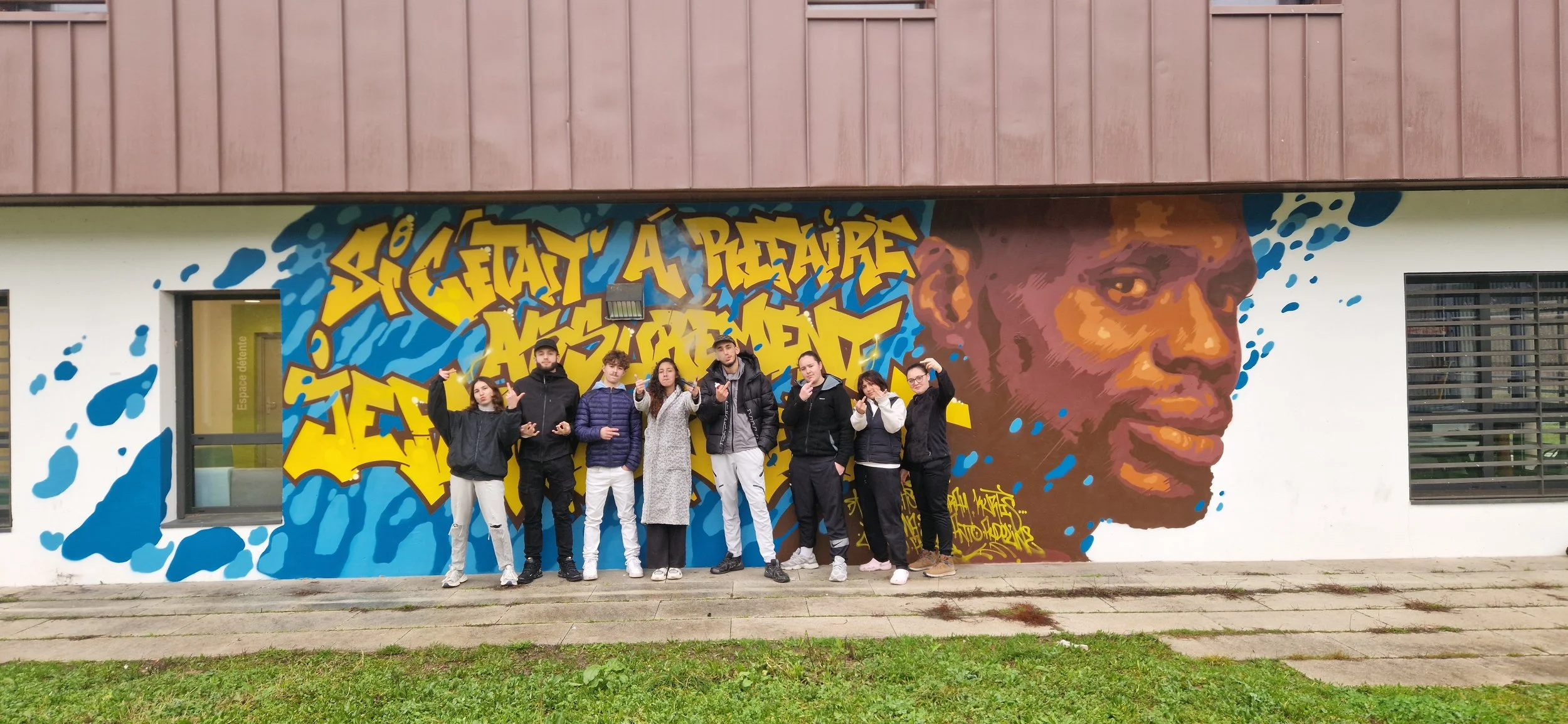 Groupe de sept jeunes debout devant une fresque murale colorée avec une tête d'homme et des graffitis