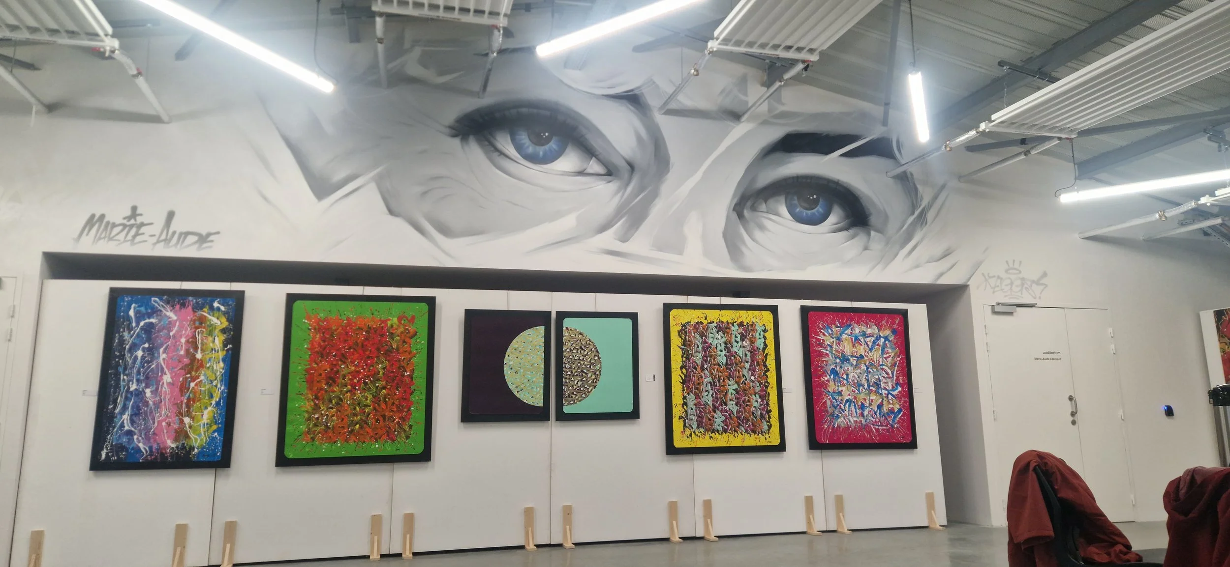 Un mur avec une fresque en noir et blanc représentant deux yeux bleus. En dessous, plusieurs tableaux d'art abstrait coloré sont exposés.