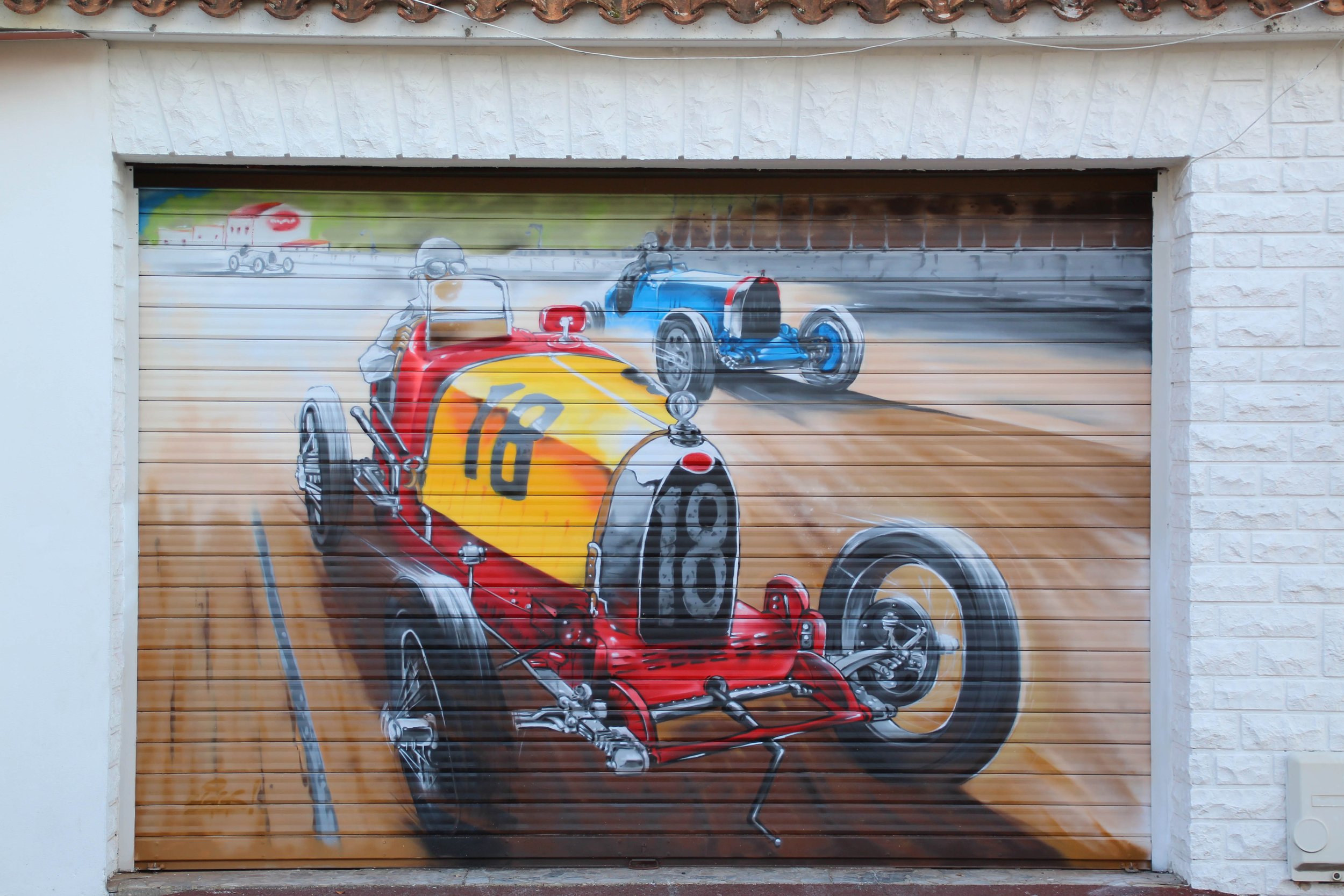Peinture murale représentant une course de voitures vintage sur une porte de garage, avec deux voitures en premier plan, une rouge et une bleue, et d'autres voitures en arrière-plan.