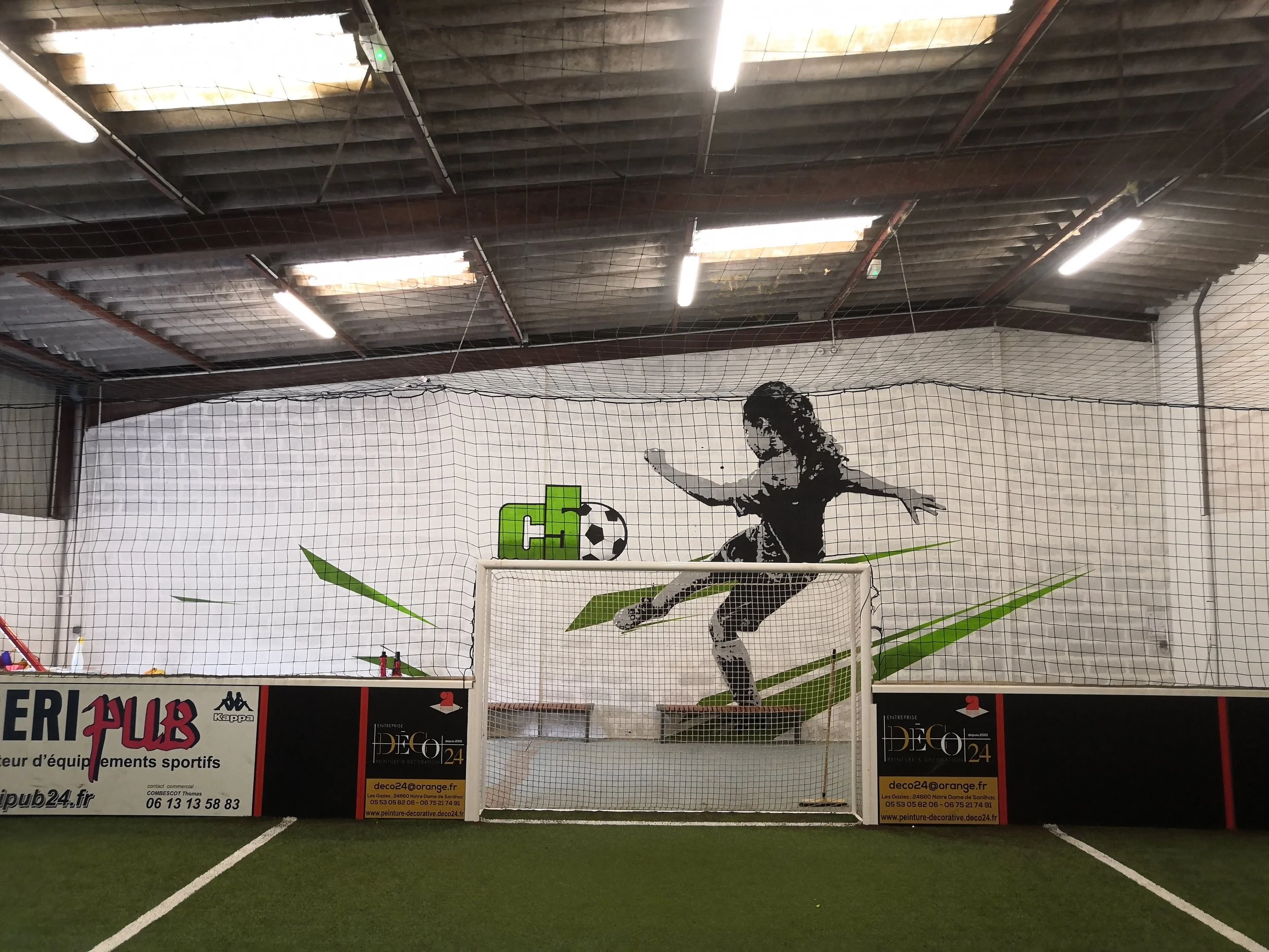 Une salle d'intérieur avec un filet de sport séparant deux zones, une affiche de sport avec un joueur en train de faire un saut pour frapper un ballon, au fond un plafond en bois avec des lumières suspendues, et un vestibule avec des panneaux publicitaires.