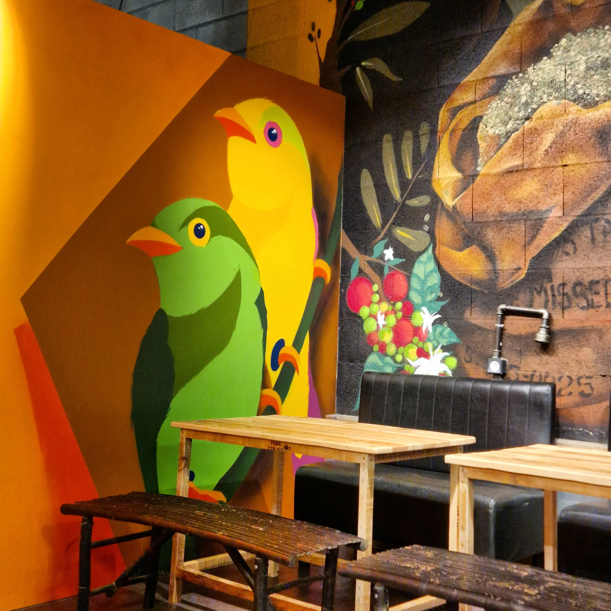 Une peinture murale colorée représentant deux oiseaux, un vert et un jaune, dans un cadre intérieur avec des meubles en bois et un sol en pierre.