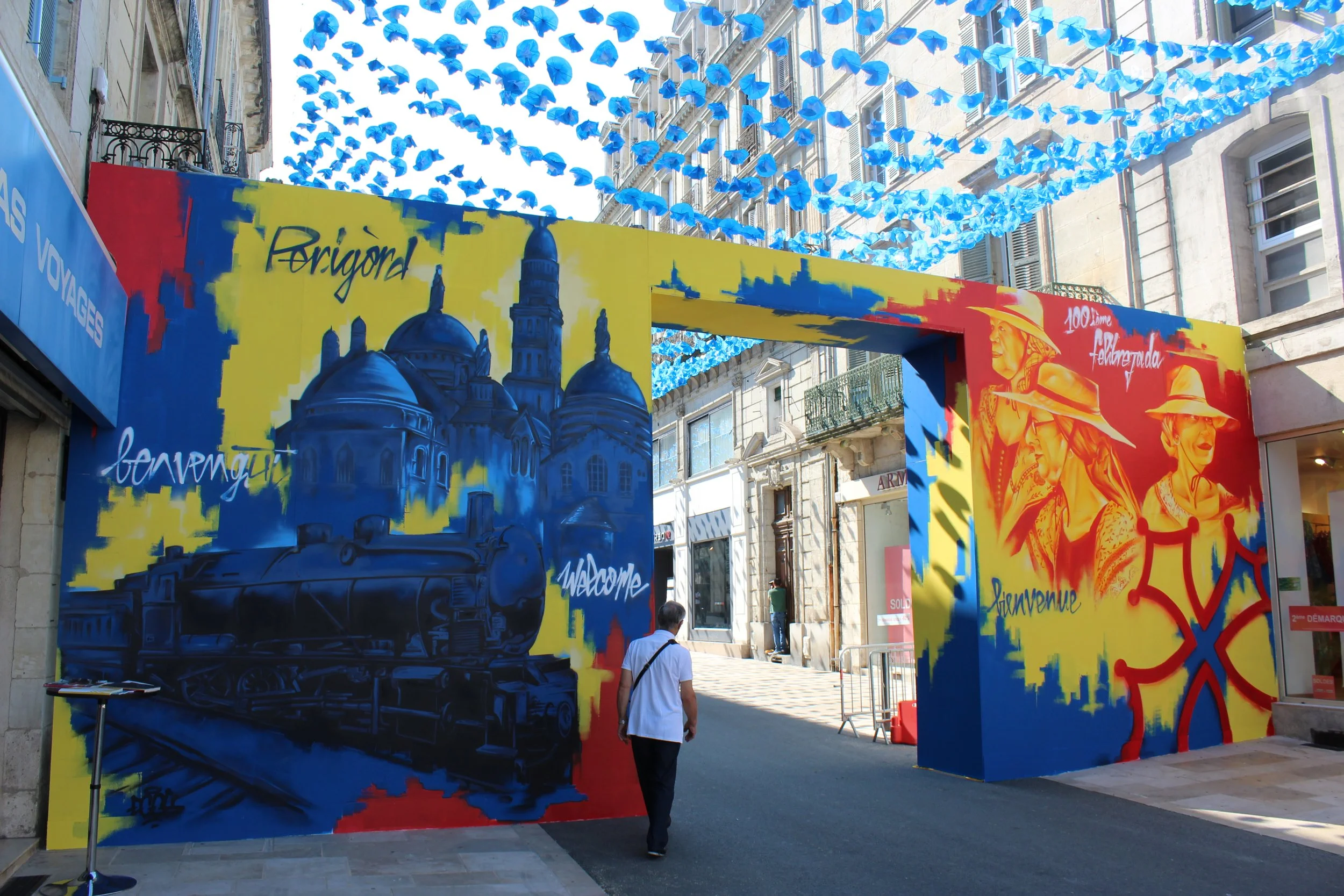 Une installation artistique urbaine avec des peintures murales colorées, représentant un train, l'eglise st front , et des figures humaines, sous un ciel décoré de rubans bleus suspendus. Périgueux Félibree porte