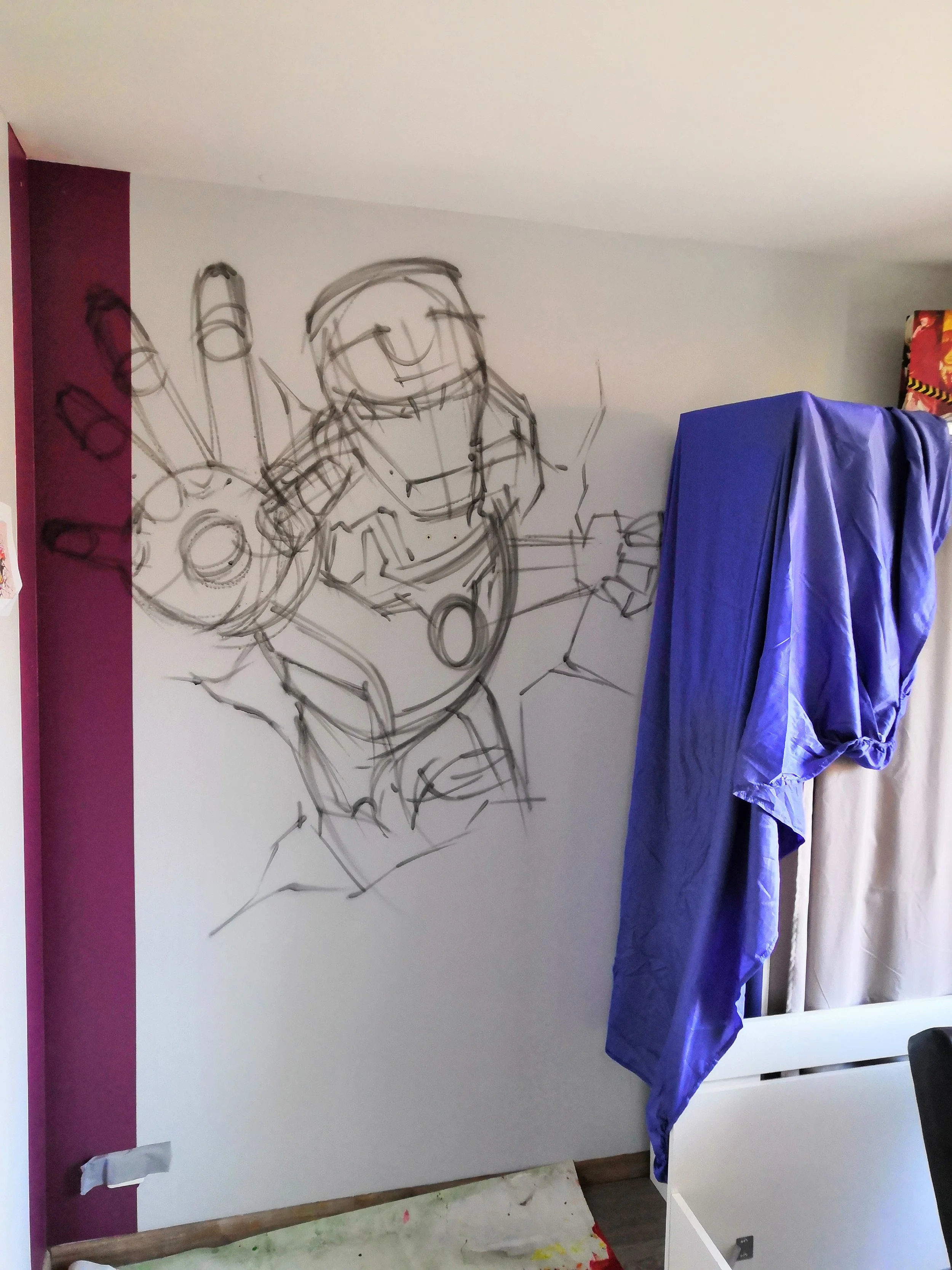 Un mur blanc avec une fresque dessinée à la main représentant un personnage souriant. Il y a un meuble ou un objet recouvert d'un drap violet à droite.