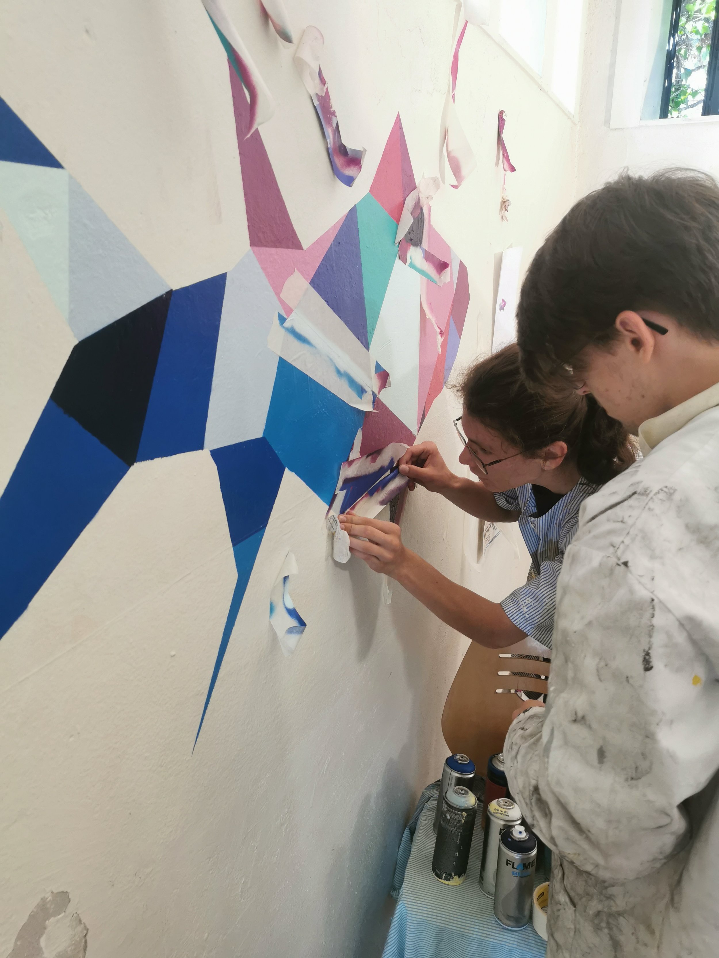 Deux personnes peignent un mur avec des formes géométriques en couleurs pastel. Plusieurs aérosols de peinture sont posés sur une surface en dessous. EGGS EGGS58STUDIO