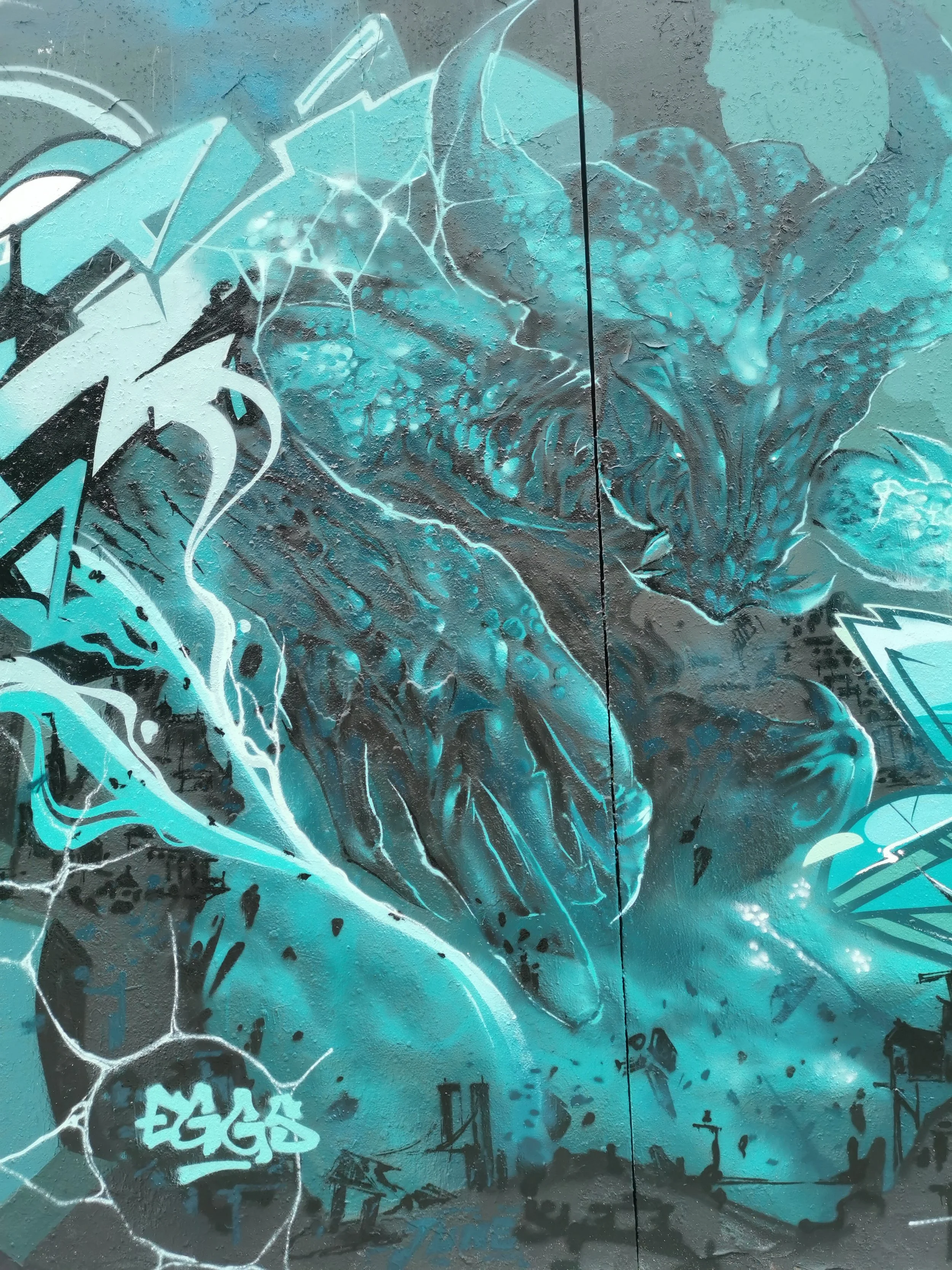 Graffiti abstrait en tons de bleu, noir et blanc sur un mur en béton, avec des formes géométriques et des lettres.