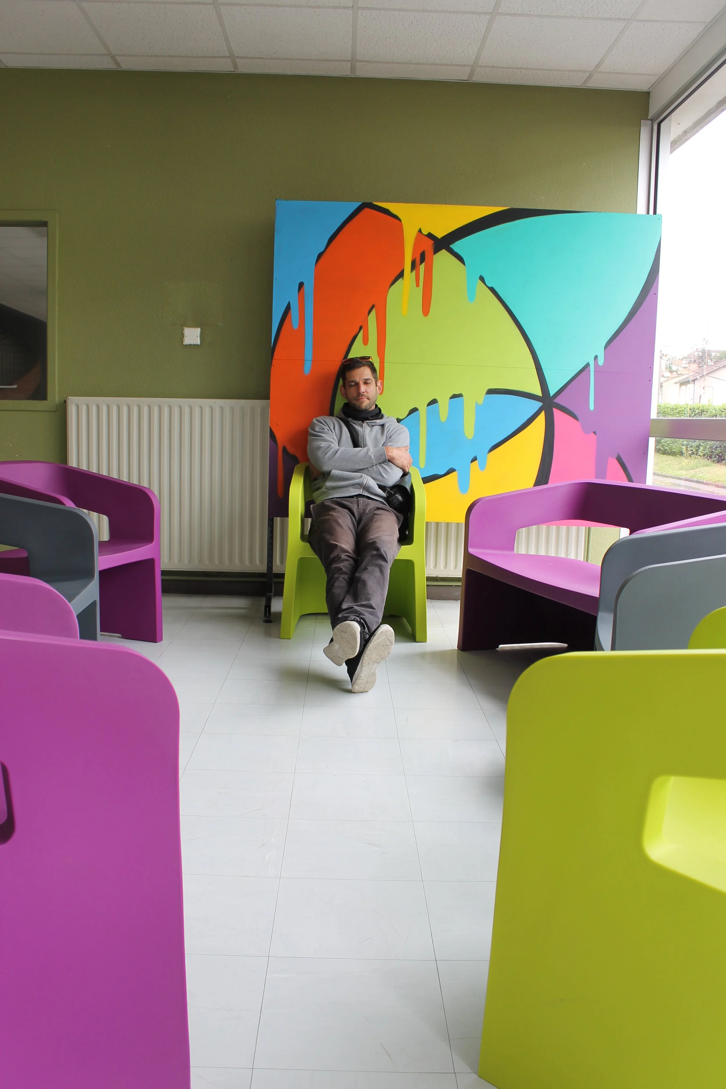 Eggs assis dans une pièce colorée, avec un fond de peinture murale abstraite aux couleurs vives, entouré de chaises violet, gris, et vert lime.
