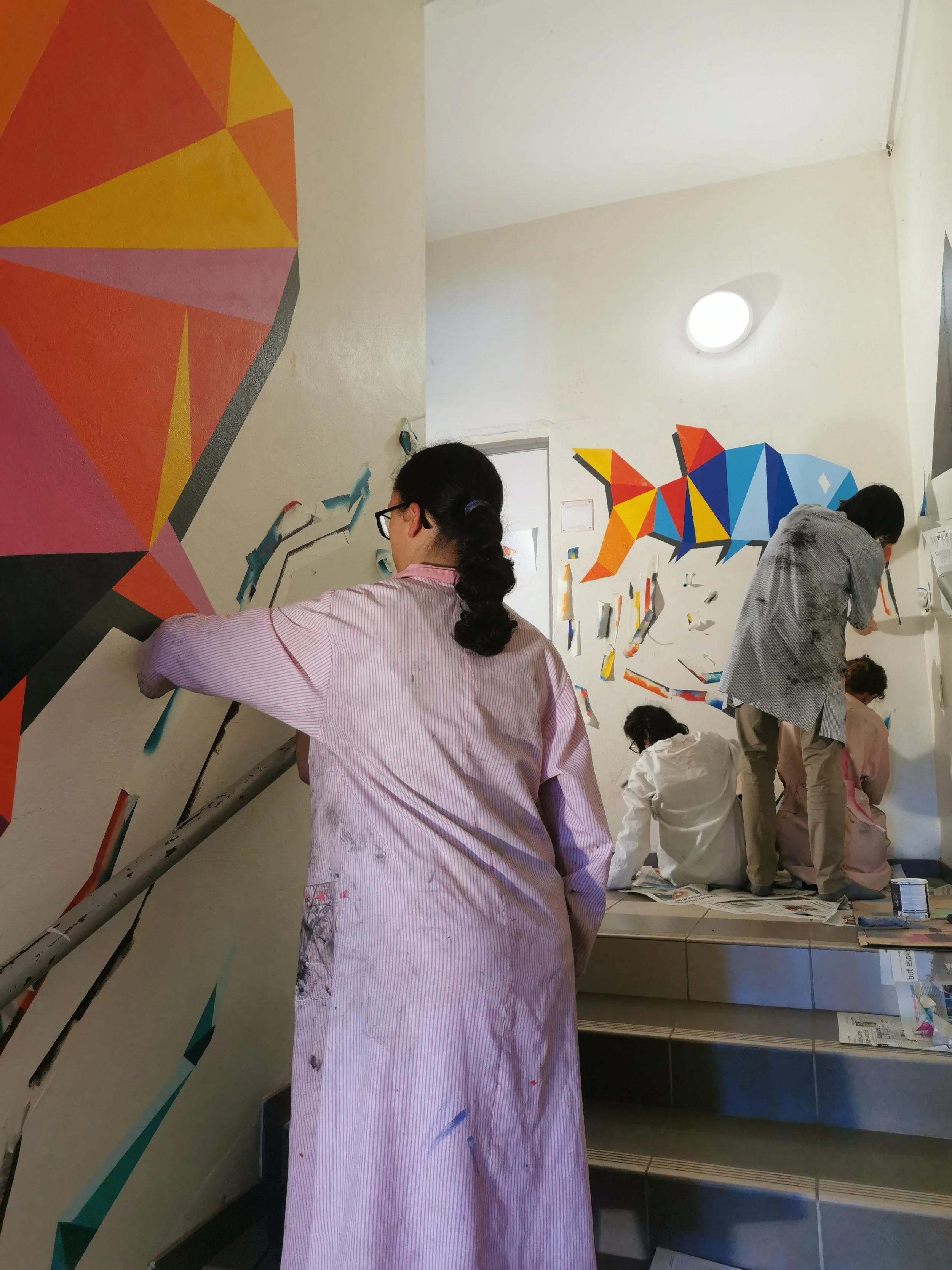 Des personnes peignent une fresque géométrique colorée sur un mur intérieur, en utilisant des pinceaux et de la peinture. EGGS EGGS58STUDIO