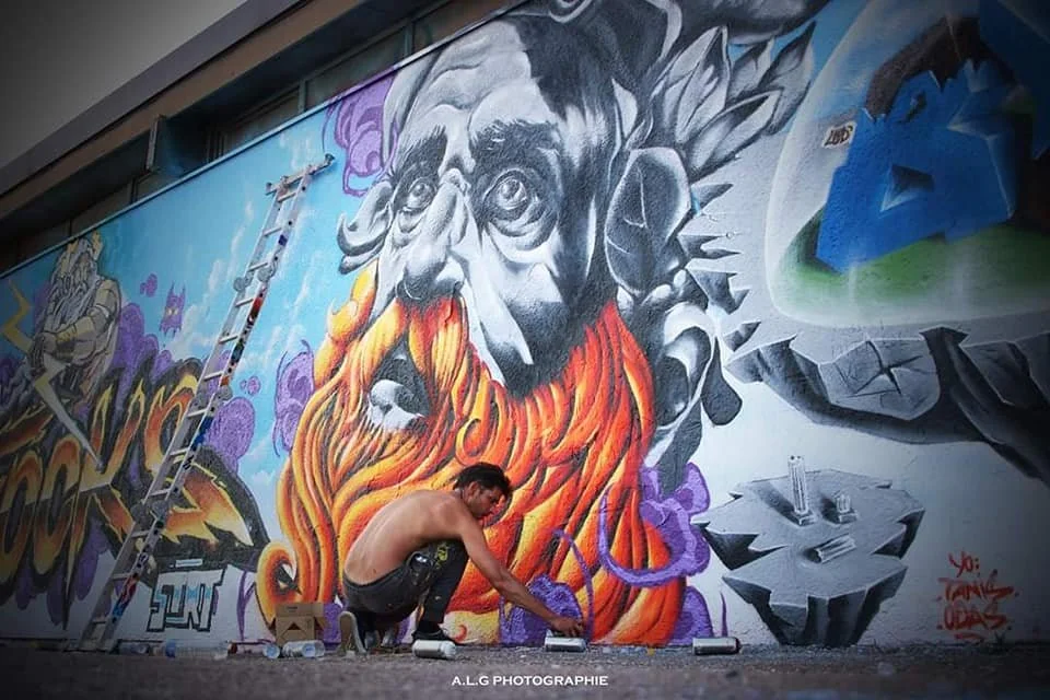 Un artiste de graffiti en train de peindre un mural détaillé représentant une figure avec une longue barbe en flammes et un visage d'homme âgé, sur un mur extérieur urbain.