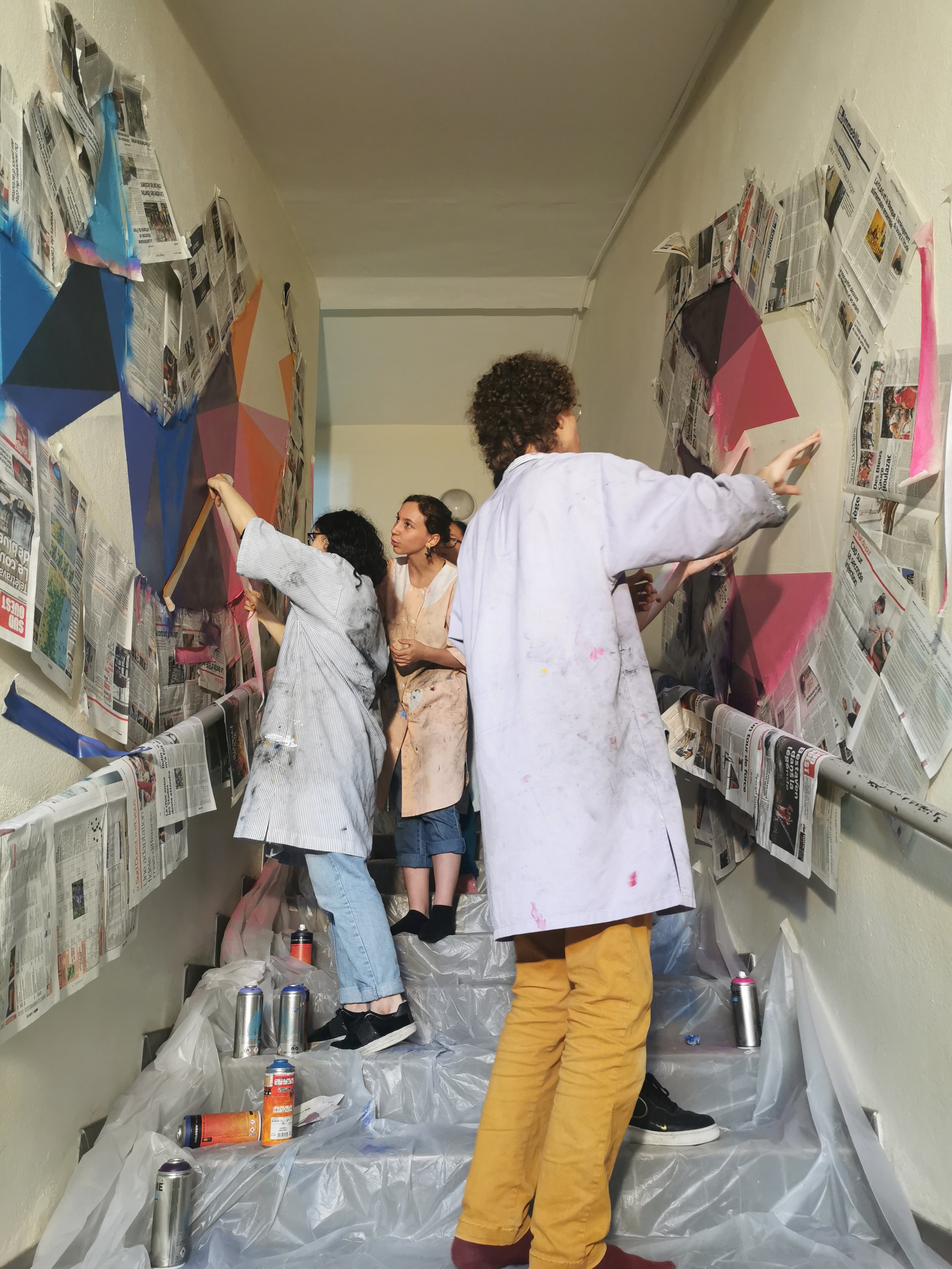 Groupe de personnes, certains peintres, en train de peindre une fresque ou mur en utilisant de la peinture acrylique et des pinceaux, dans un espace couvert avec des murs tapissés de journaux. EGGS EGGS58STUDIO