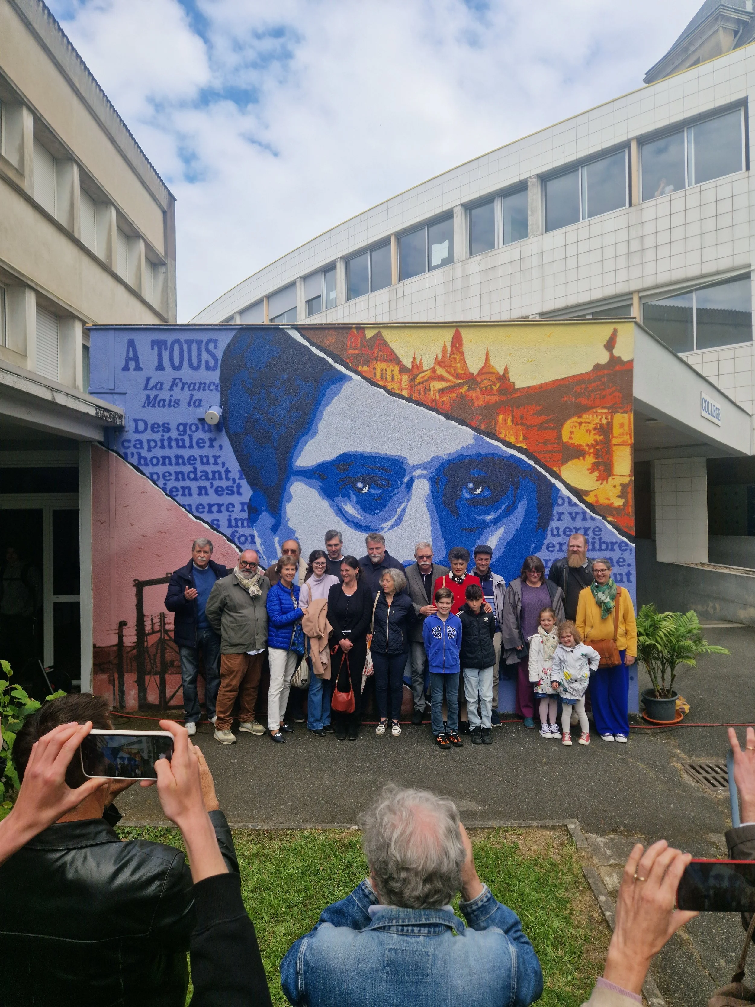 Groupe de personnes posant devant une fresque murale de EGGS EGGS58STUDIO représentant une personne portant des lunettes, avec un fond de ville historique. Plusieurs personnes prennent des photos ou regardent vers le groupe. Laure Gatet