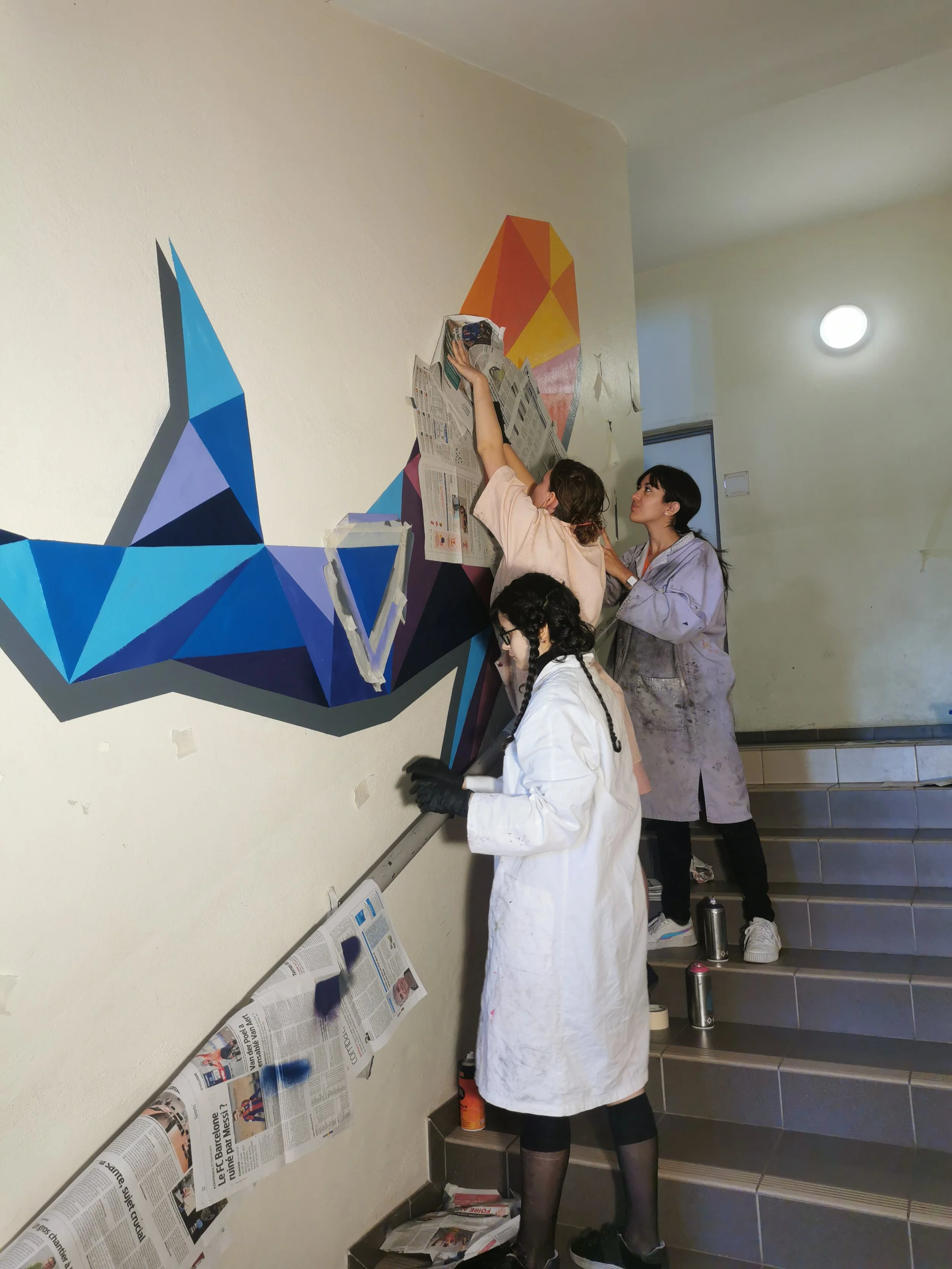Des personnes en train de décorer un mur avec un motif géométrique coloré en utilisant de la colle ou de la peinture. Une femme en blouse blanche et en leggings noirs travaille à fixer des journaux sur le mur, tandis qu'une autre femme en vêtement gr
