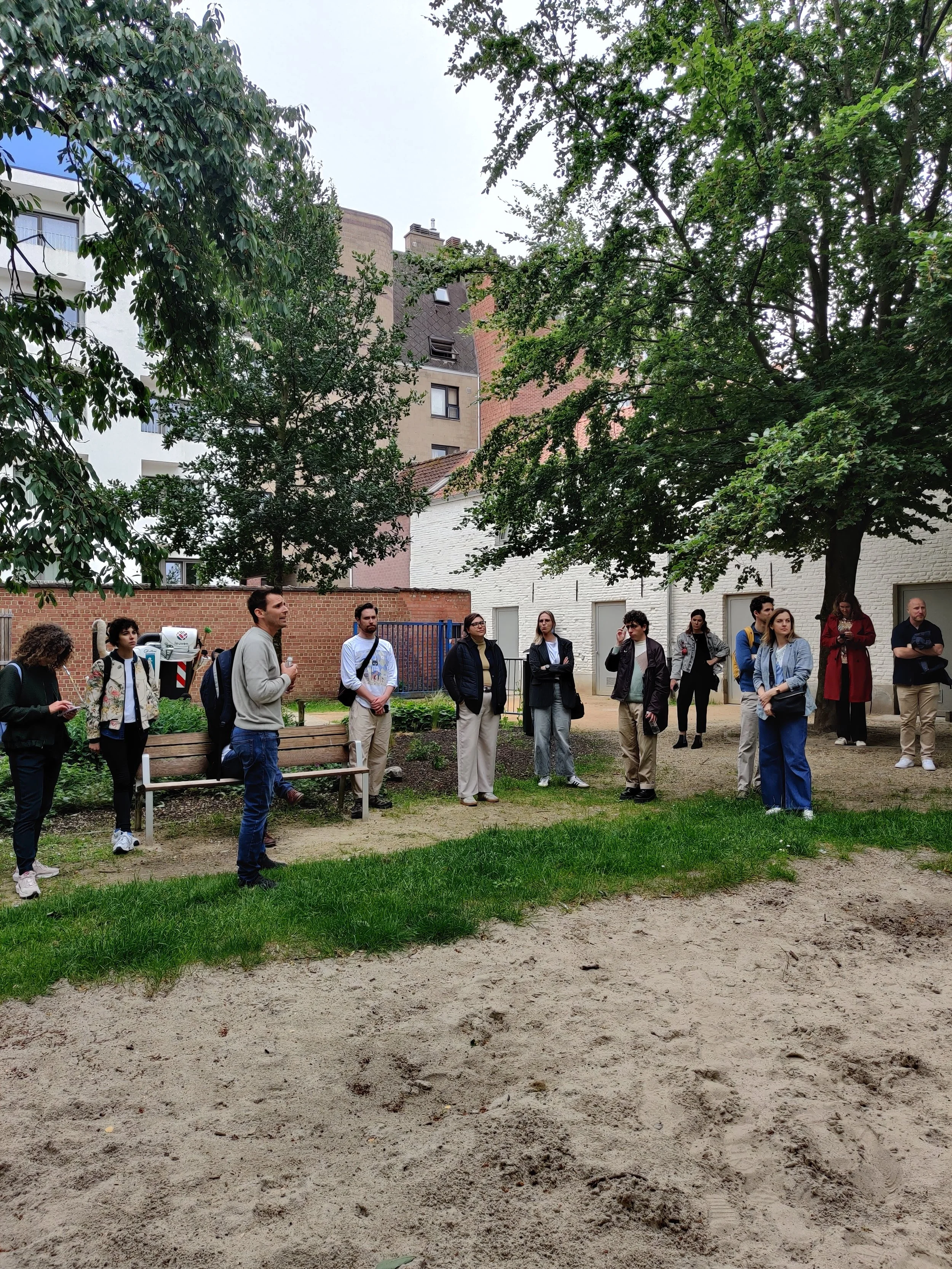 A group of people gathered outdoors under a large tree with buildings in the background, some standing and observing, others talking, on a partly cloudy day.