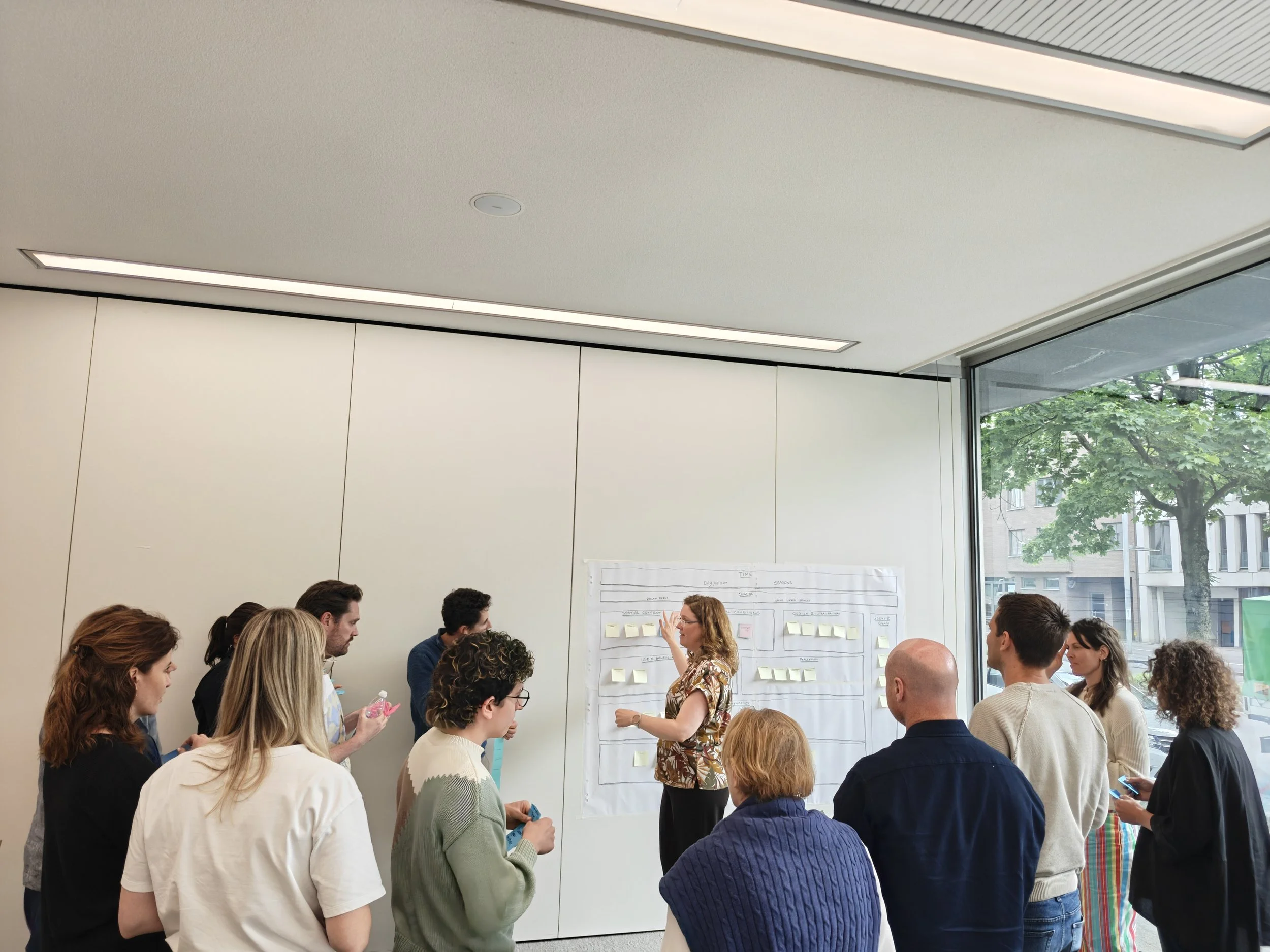 A group of people gathered in a modern conference room, listening to a woman presenting a large poster with notes and diagrams. Large windows reveal trees outside, and the room has a high ceiling with built-in lighting.