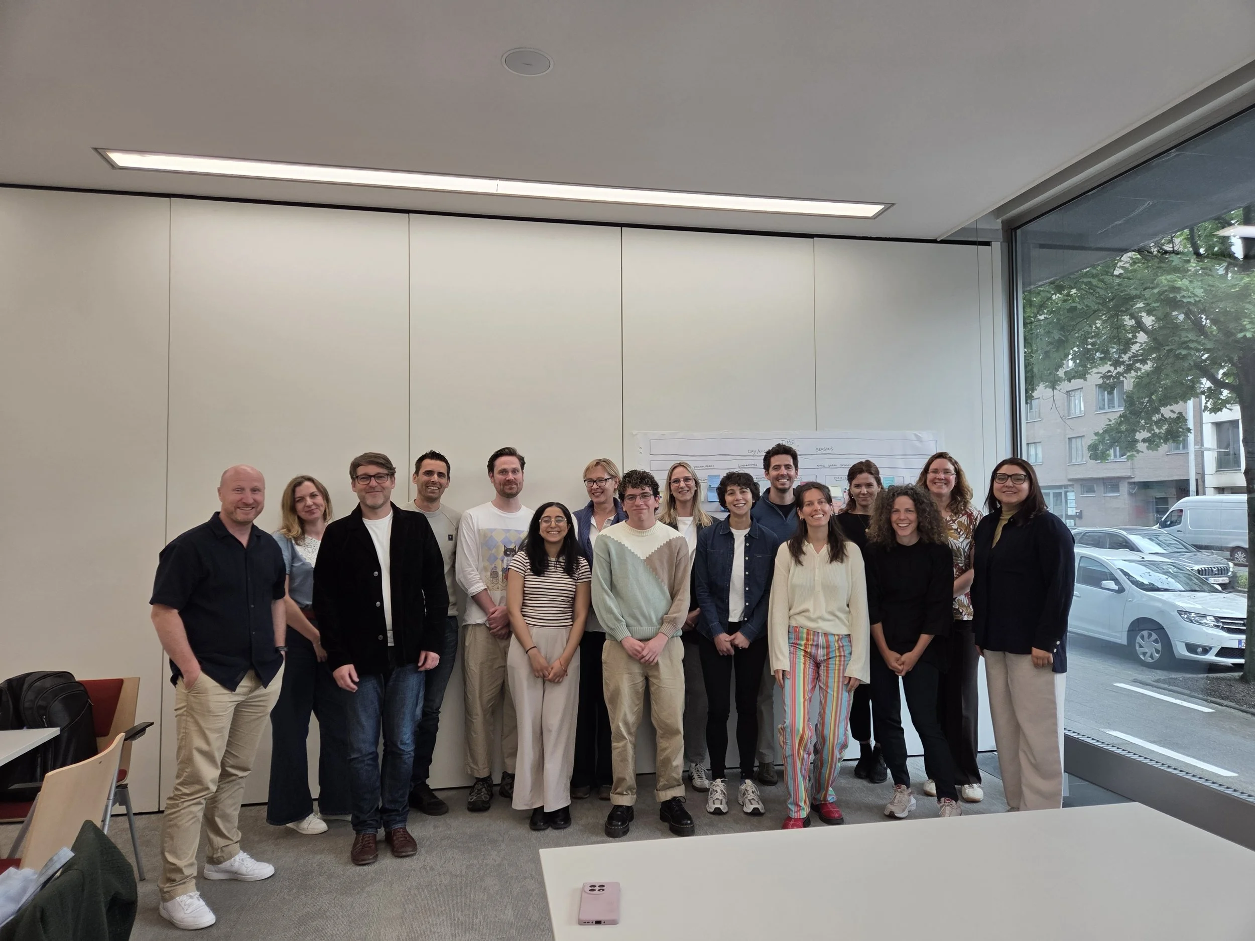 A group of 17 diverse people, smiling, standing inside a modern conference room with large windows showing trees and buildings outside. Some are wearing glasses and casual clothing, and they are posing for a group photo.