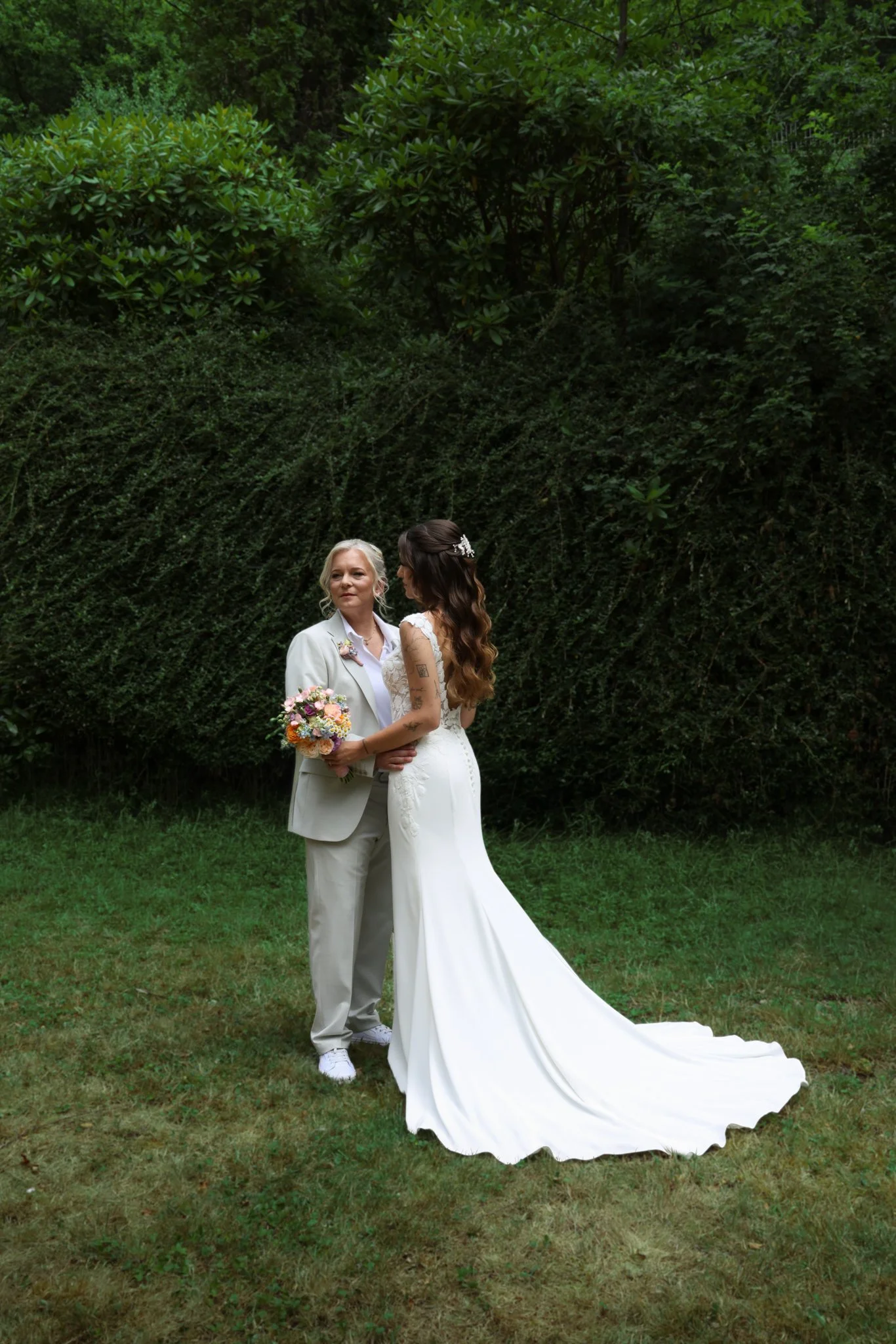 Zwei Bräute in Hochzeitskleidern stehen sich lächelnd gegenüber und halten einen Brautstrauß – authentische Hochzeitsreportage im Grünen.