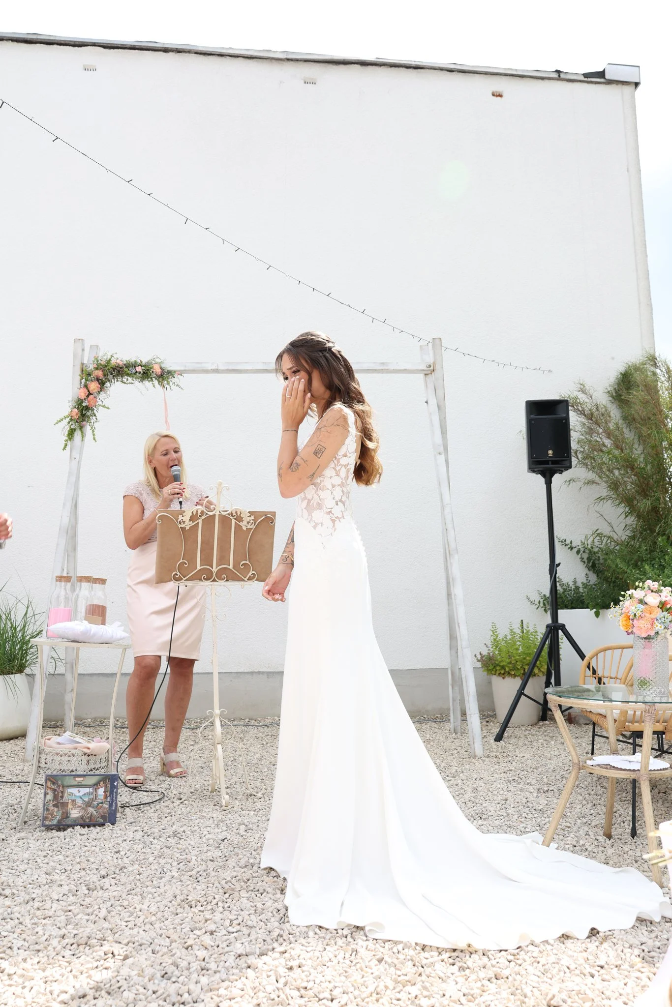 A bride in a white wedding dress appears emotional while possibly crying, holding her hand to her face, during a wedding ceremony. An officiant woman with blonde hair is speaking into a microphone and standing behind a decorative music stand. The out