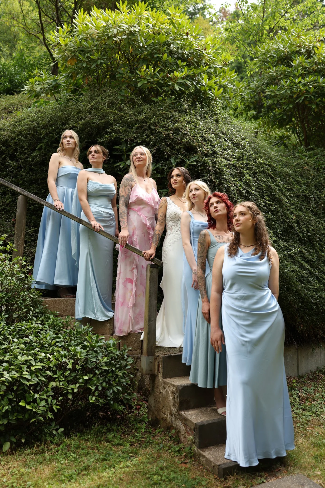 Seven women standing on a stone staircase outdoors among greenery and trees, dressed in pastel-colored formal dresses.