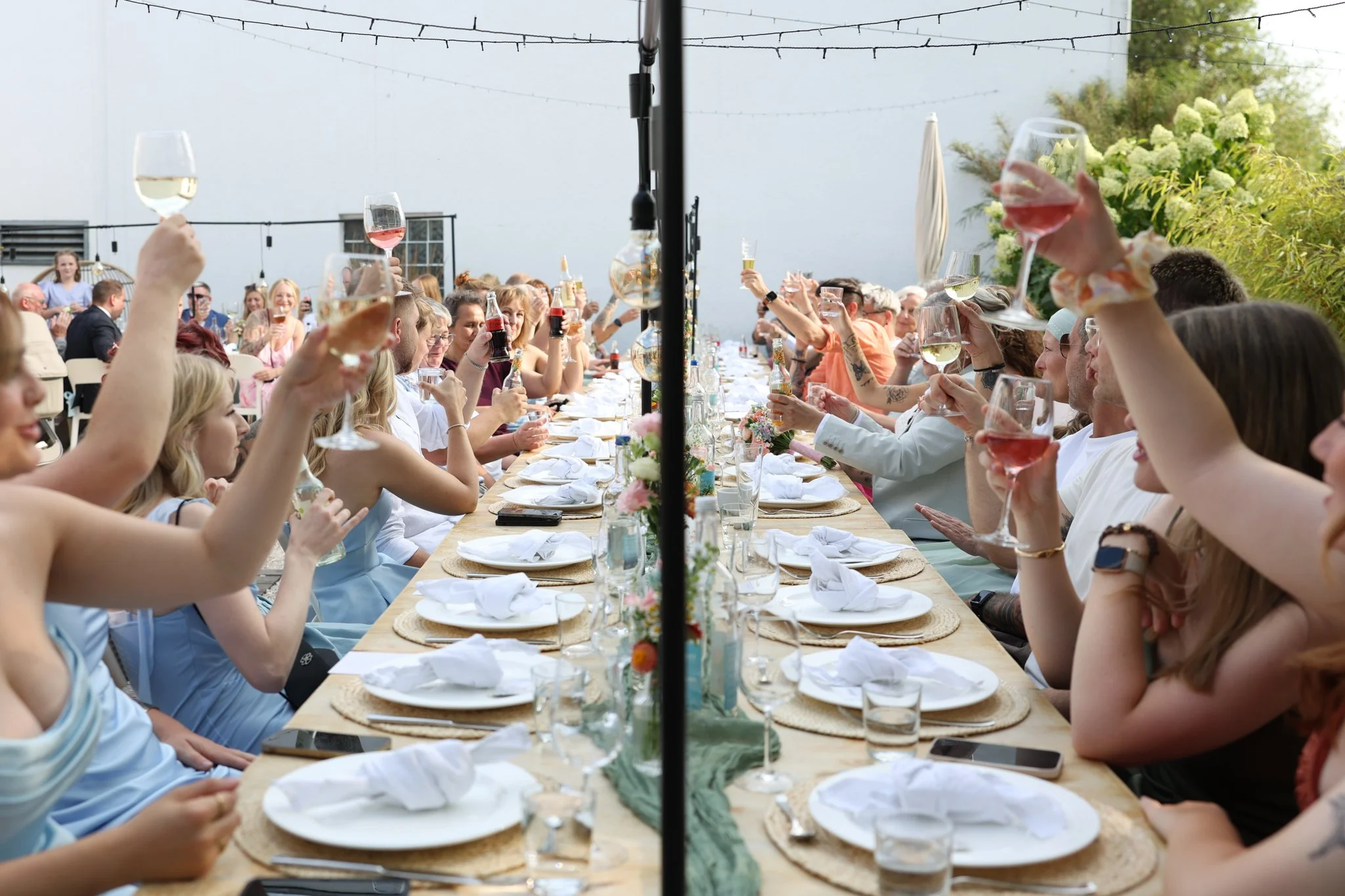 A large group of people gathered at a long outdoor dining table, raising glasses in a toast. The scene is festive, with string lights overhead and greenery in the background, suggesting a celebration or special occasion.