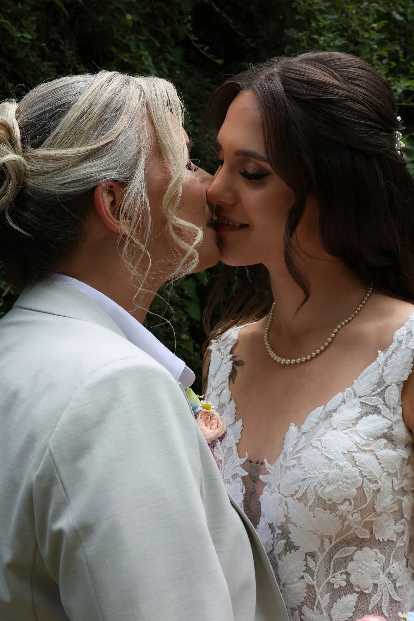 Two women in wedding attire face each other closely, about to kiss, with a background of green foliage.