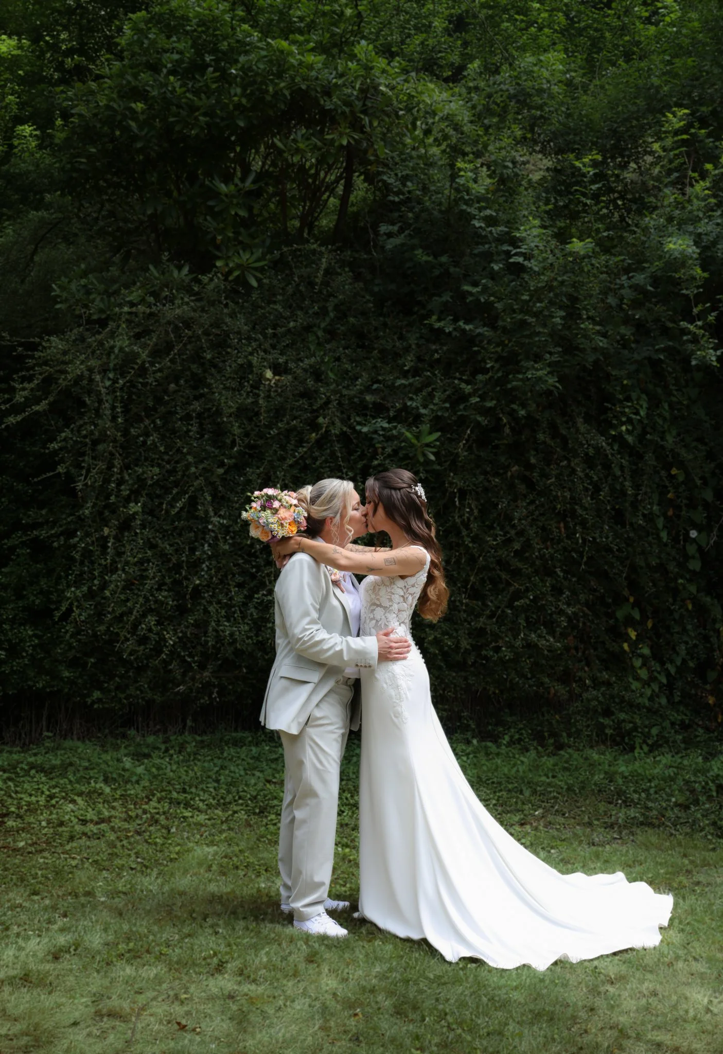 A couple dressed in wedding attire sharing a kiss outdoors, with lush green foliage in the background.