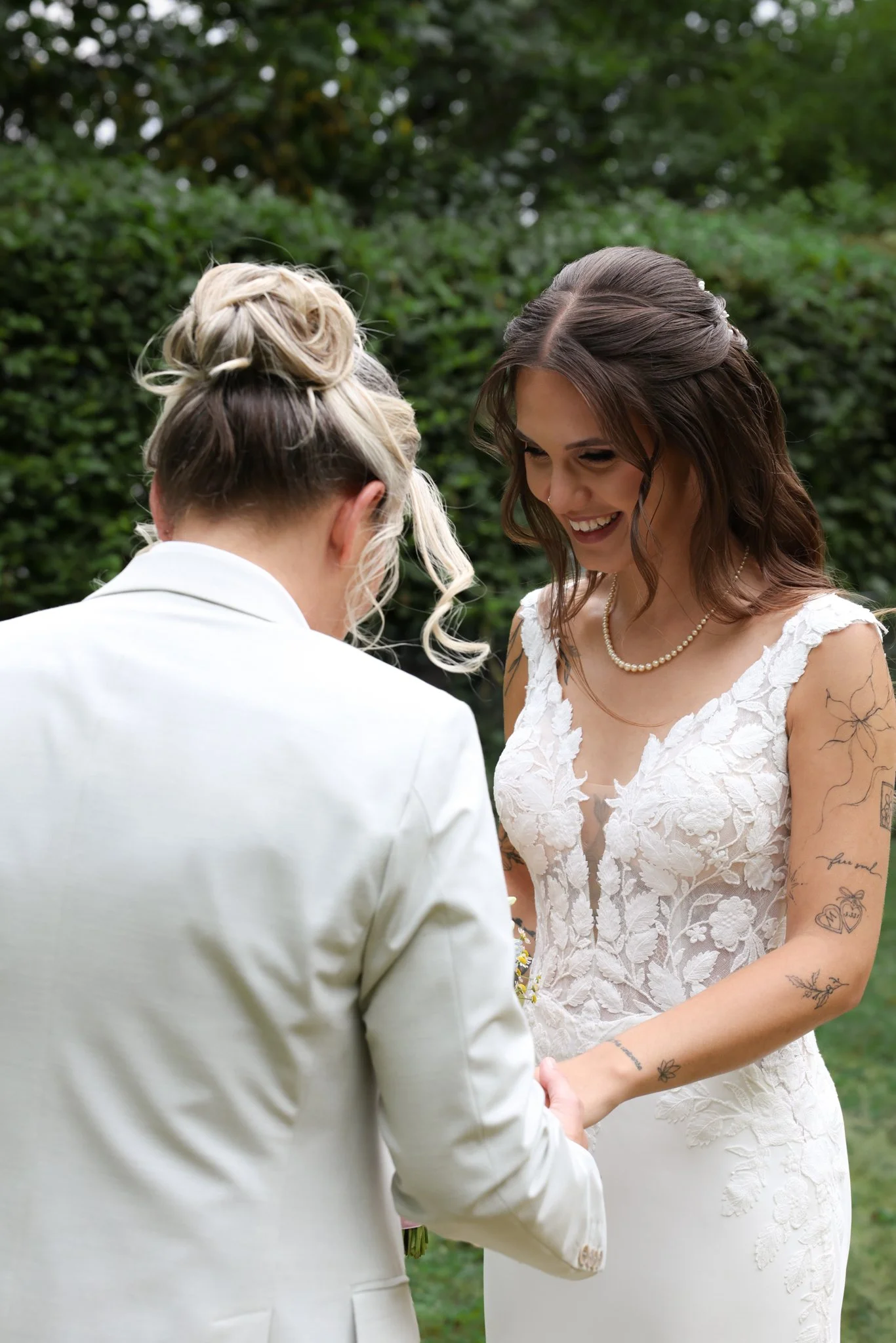 Two women holding hands during a wedding ceremony outdoors, one wearing a white wedding dress and the other in a white suit, surrounded by greenery.