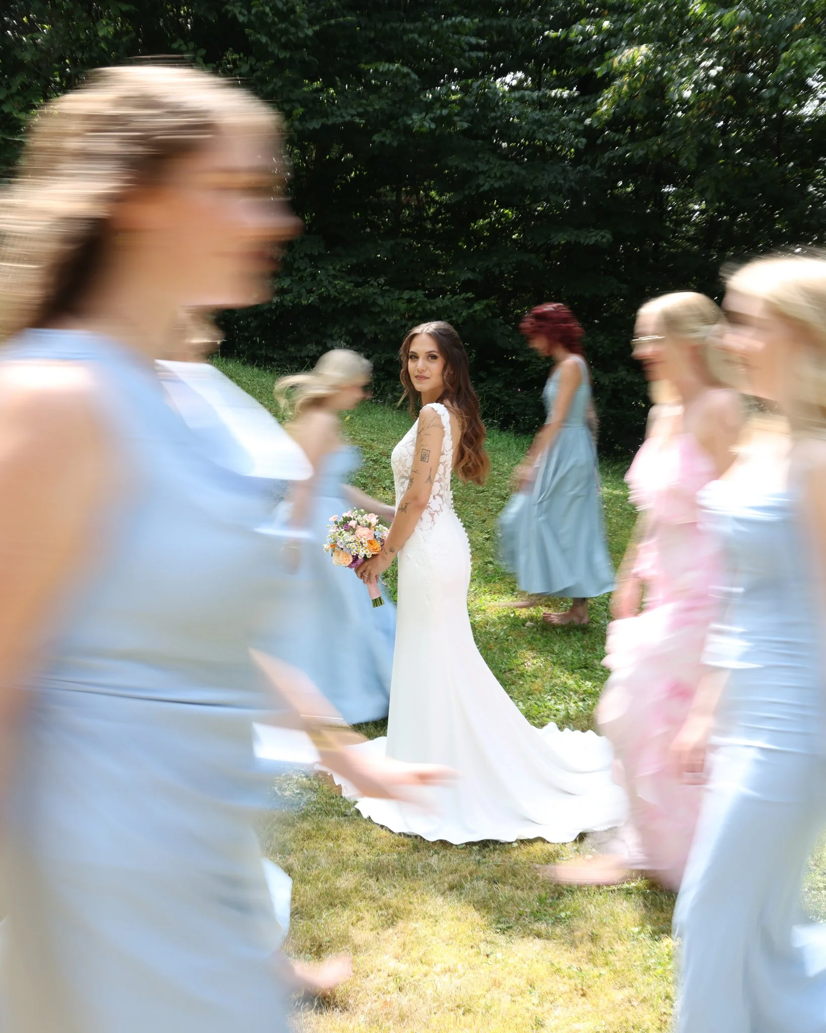 Braut im weißen Hochzeitskleid mit farbenfrohem Brautstrauß blickt in die Kamera, während sich Hochzeitsgäste im Hintergrund bewegen.